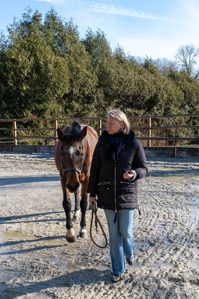 Amandine Courtois, equi coach,  avec un cheval dans un enclos en extérieur, entouré de hautes buissons et d'arbres, par une journée ensoleillée.