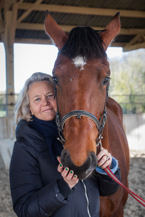 Amandine Courotis, equi-coach, porte un manteau noir et tient la tête d'un cheval marron dans une écurie.