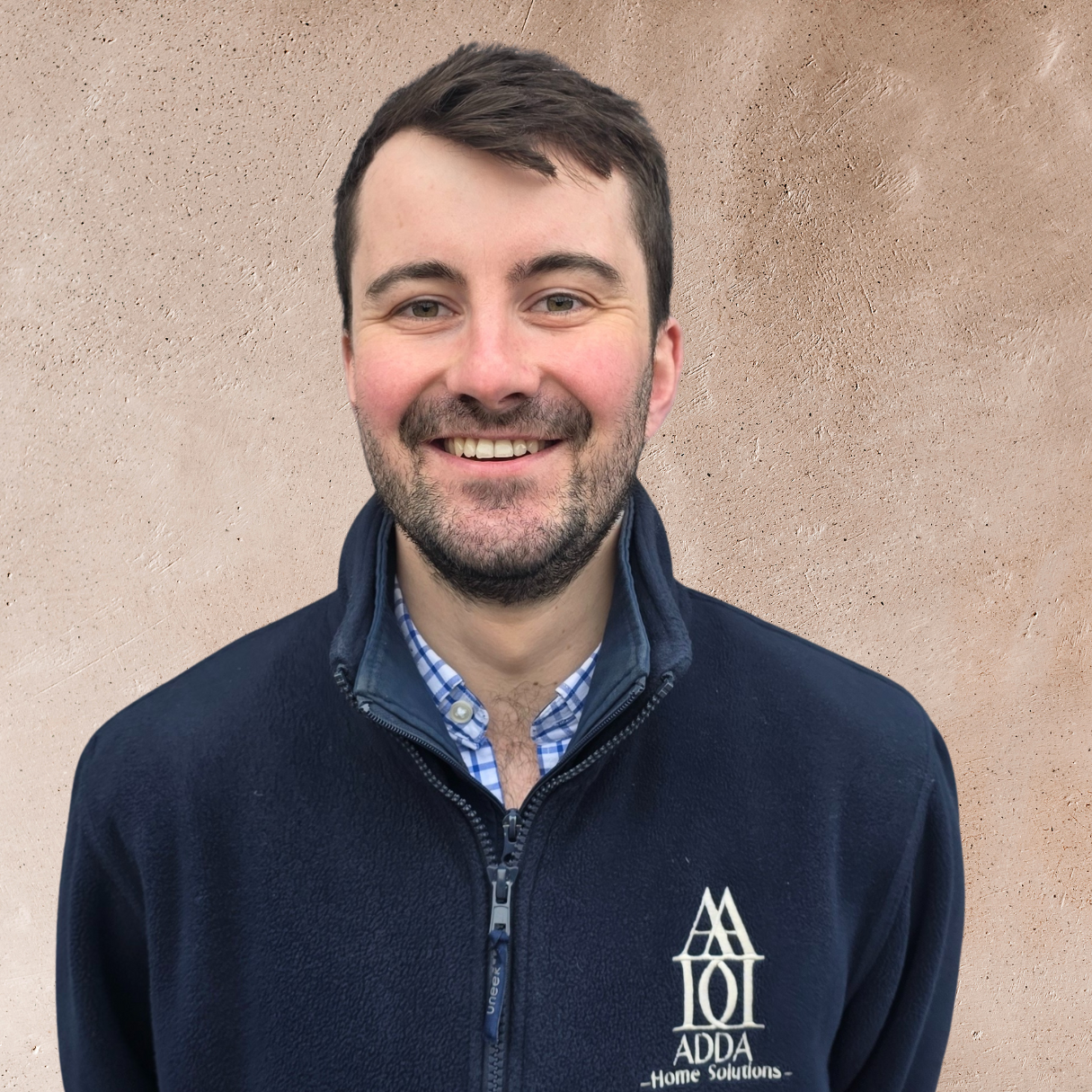 Young man with a beard and short dark hair, wearing a light blue t-shirt and navy blue vest, standing outdoors with arms crossed, smiling, in front of a wooden barrel and a brick wall, with greenery and hanging lights in the background.