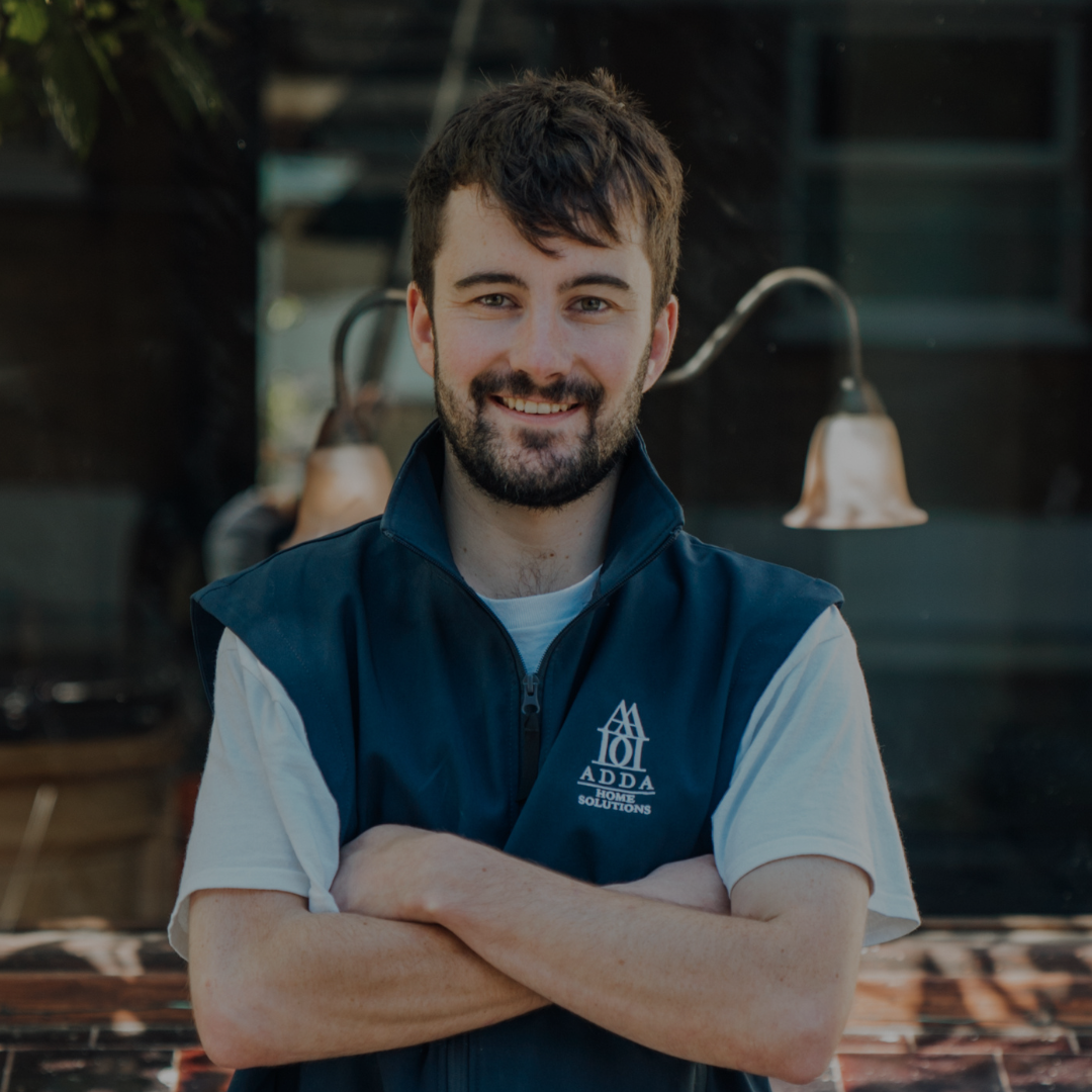 A smiling man with dark hair and beard, wearing a blue vest with a logo, standing outdoors with arms crossed in front of a storefront with outdoor lighting.