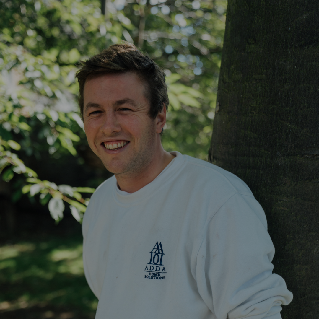 A young man smiling and leaning against a tree outdoors, wearing a white sweatshirt with a logo that reads 'ADDA Home Solutions'.