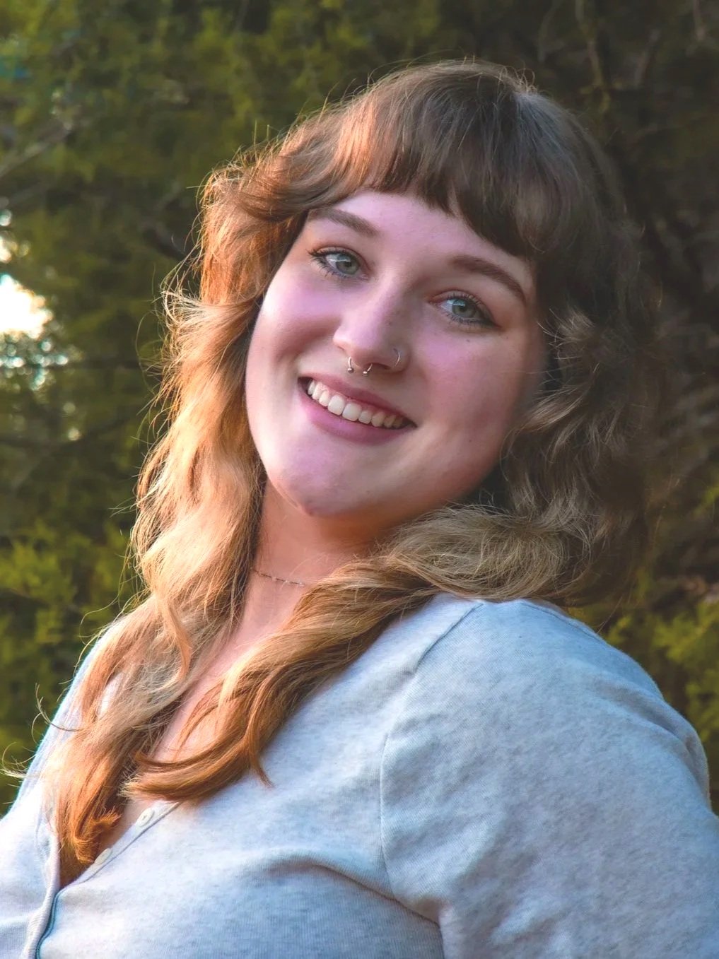 A woman with brown, wavy hair and blue eyes smiling outdoors in front of green trees.