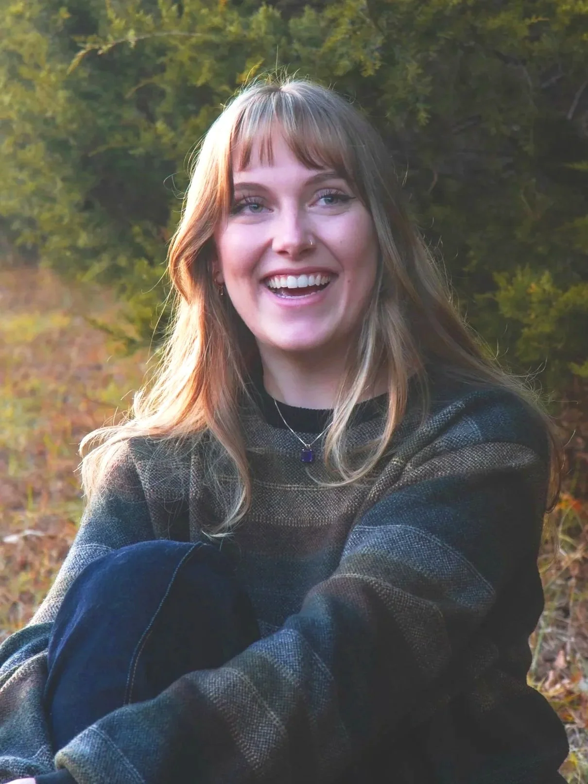 A young woman with blonde hair and bangs smiling outdoors in a forested area during daylight.