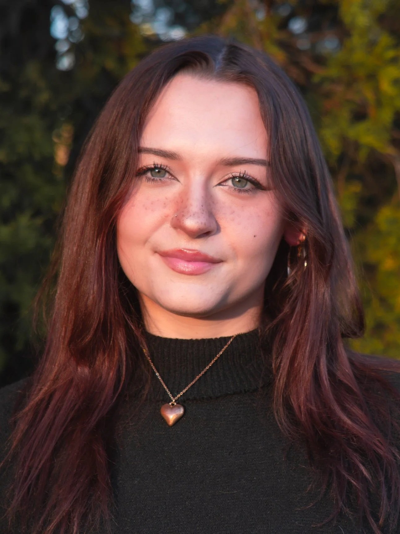 Close-up of a young woman with shoulder-length reddish-brown hair, green eyes, wearing a black turtleneck and a necklace with a heart-shaped pendant, outdoors in natural light with blurred green foliage background.