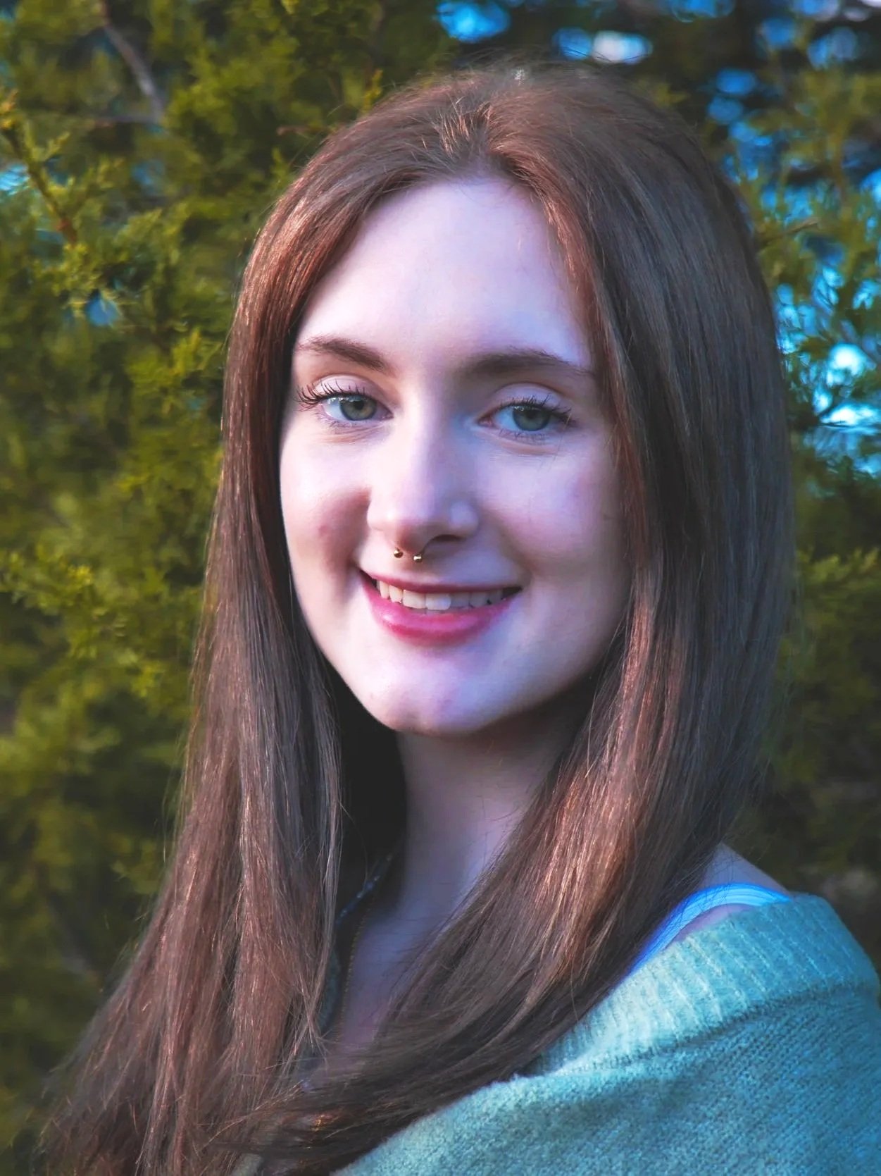 A young woman with long brown hair, light skin, and blue eyes smiling outdoors with a background of green trees.