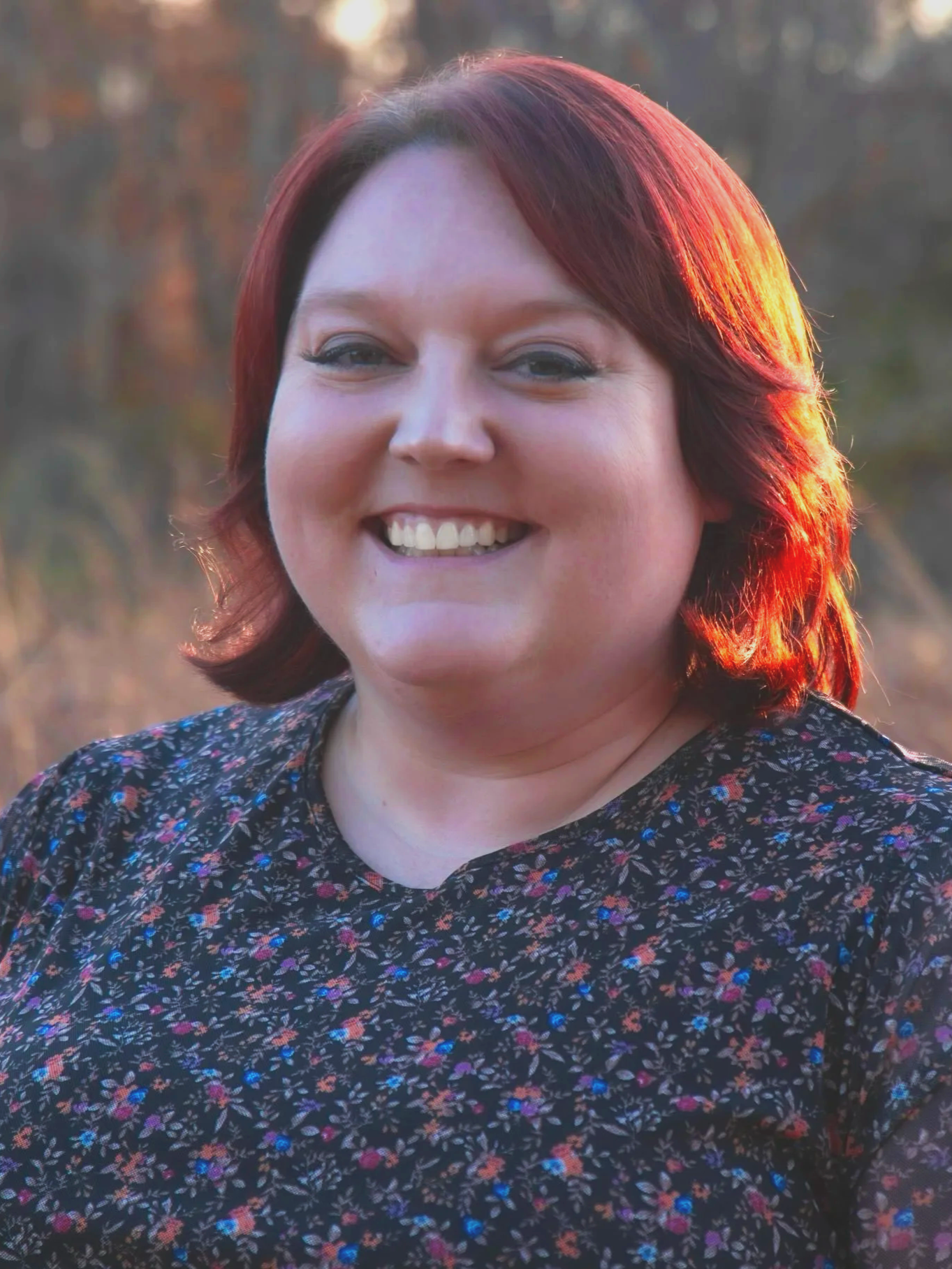 A woman with red hair smiling outdoors at sunset, wearing a dark floral patterned top.