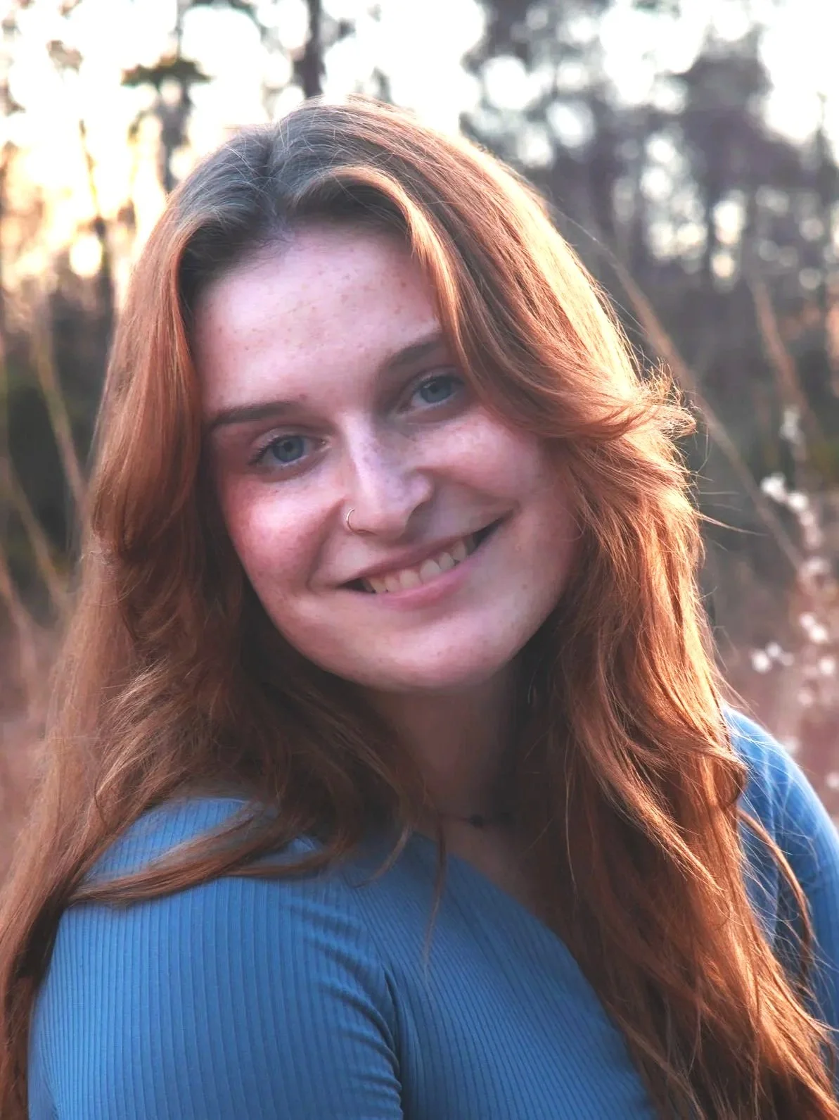 Young woman with red hair smiling outdoors at sunset, wearing a blue top, with a background of trees and blurred nature.