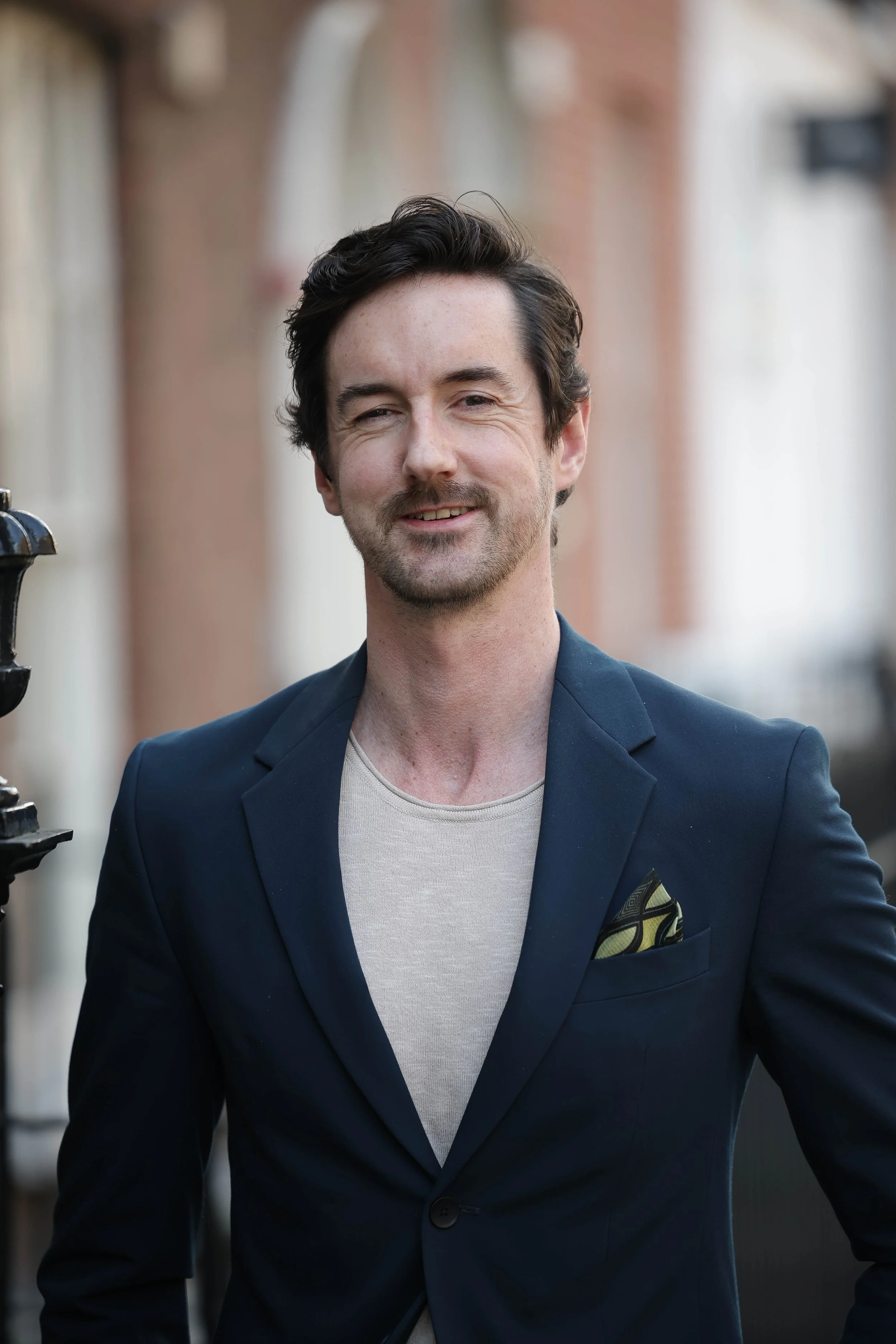 A man with dark hair and a beard, wearing a dark blue blazer with a pocket square and a light-colored t-shirt, standing outdoors in front of a brick building with blurred background.