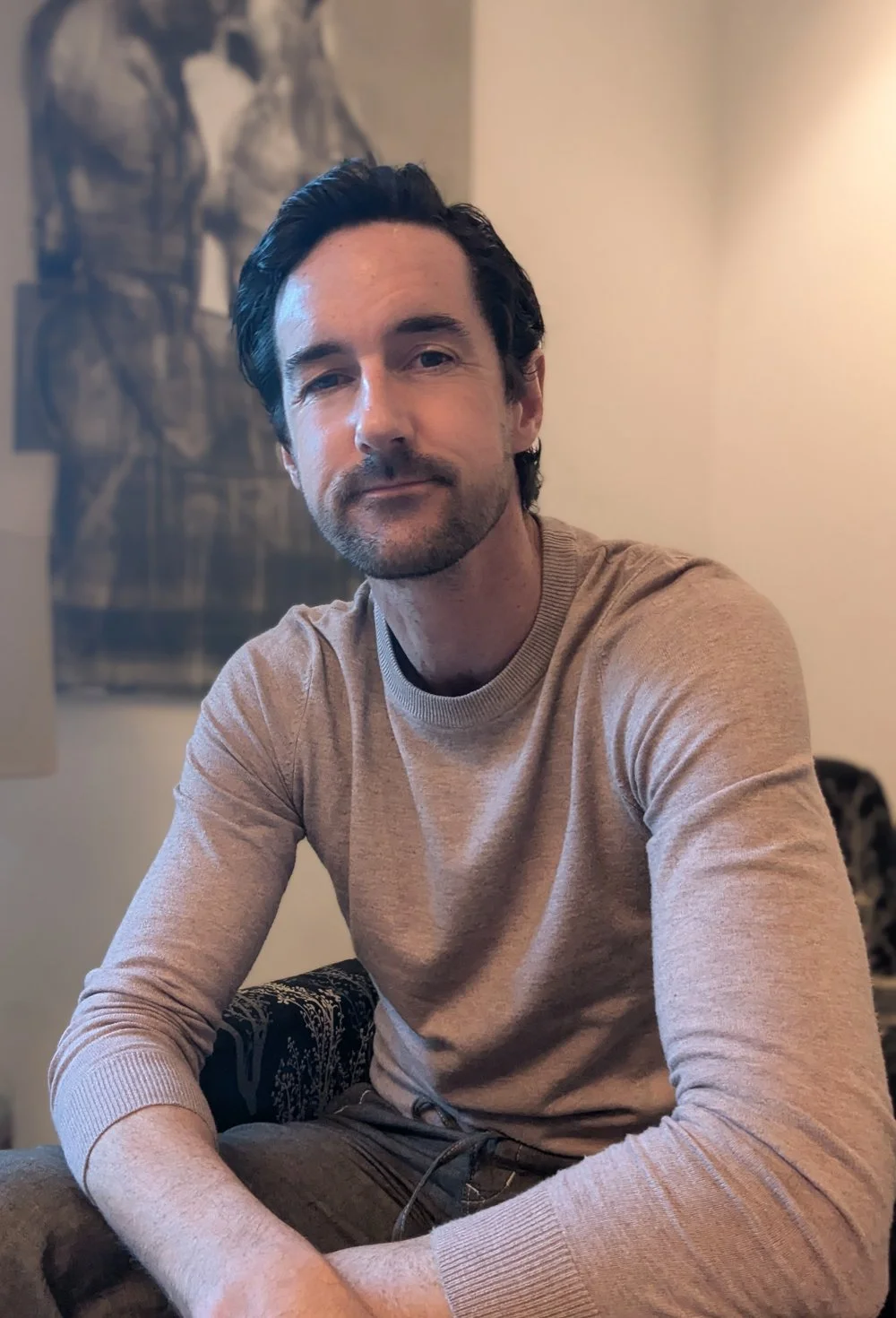 A man with dark hair and a beard sitting on a chair, wearing a beige long-sleeve shirt, in a cozy room with beige walls and artwork in the background.