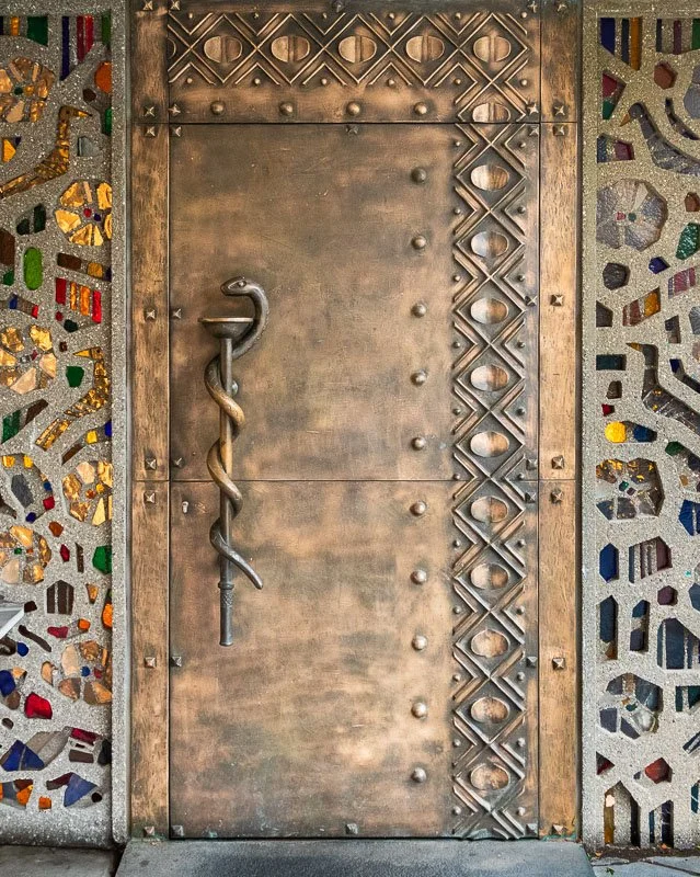 Eingangstür zur Praxis Dr. Giesen mit einem Äskulapstab als Türgriff, flankiert von filigranen, mosaikähnlichen Mustern auf beiden Seiten.