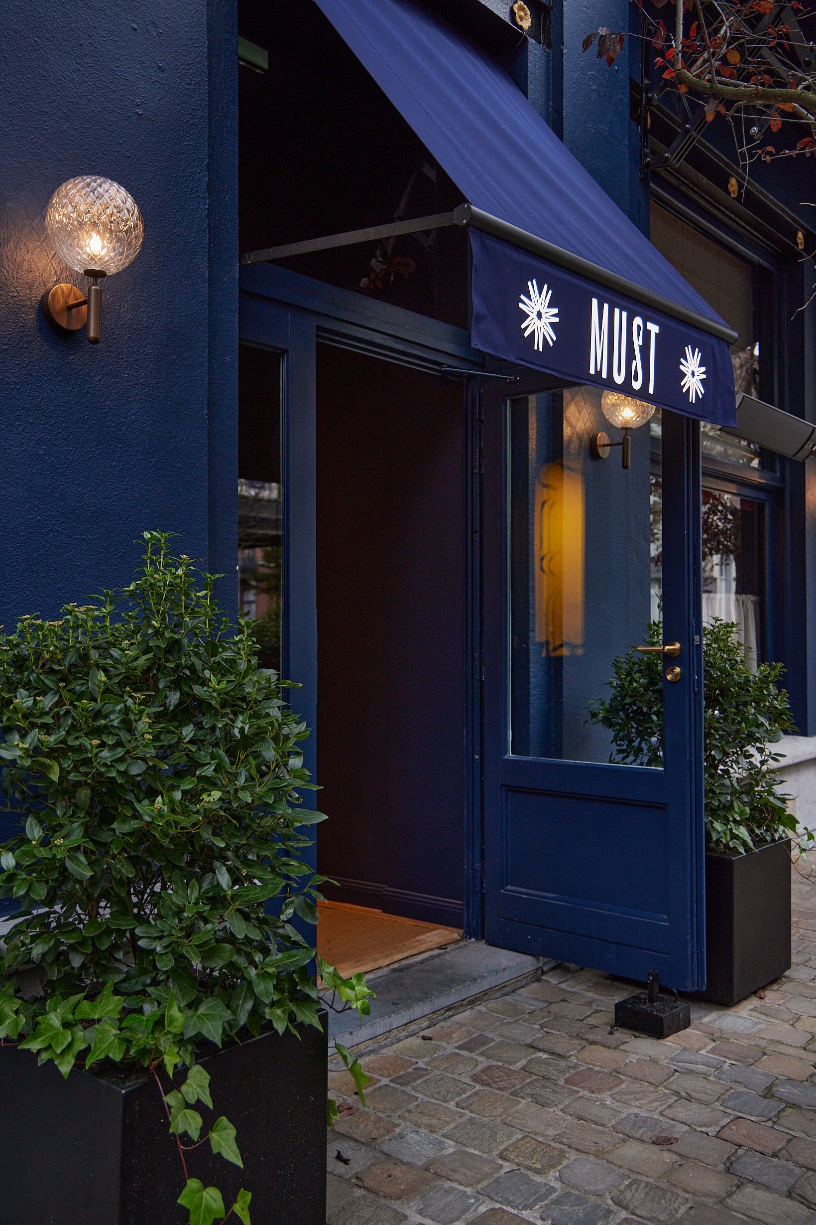 MANOS Build & Maintenance MUST restaurant working site
Entrance to a restaurant or cafe called 'MUST' with blue walls, glass door, outdoor plants in black planters, and brick pavement.