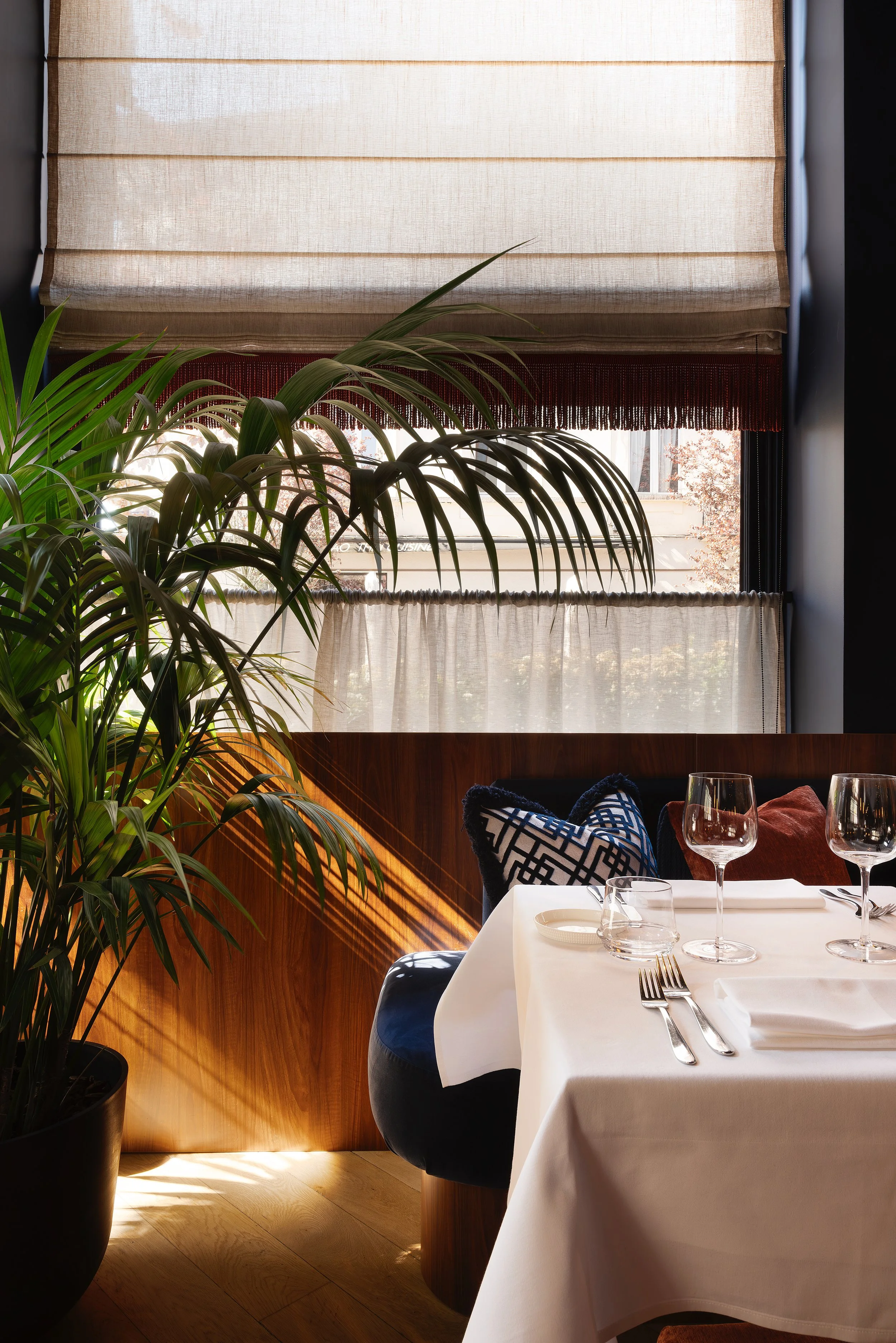MANOS Build & Maintenance MUST restaurant working site
A cozy restaurant corner with a large potted plant near a window, sunlight casting shadows on a white tablecloth, set with wine glasses, utensils, and napkins, and a decorative pillow on a bench.