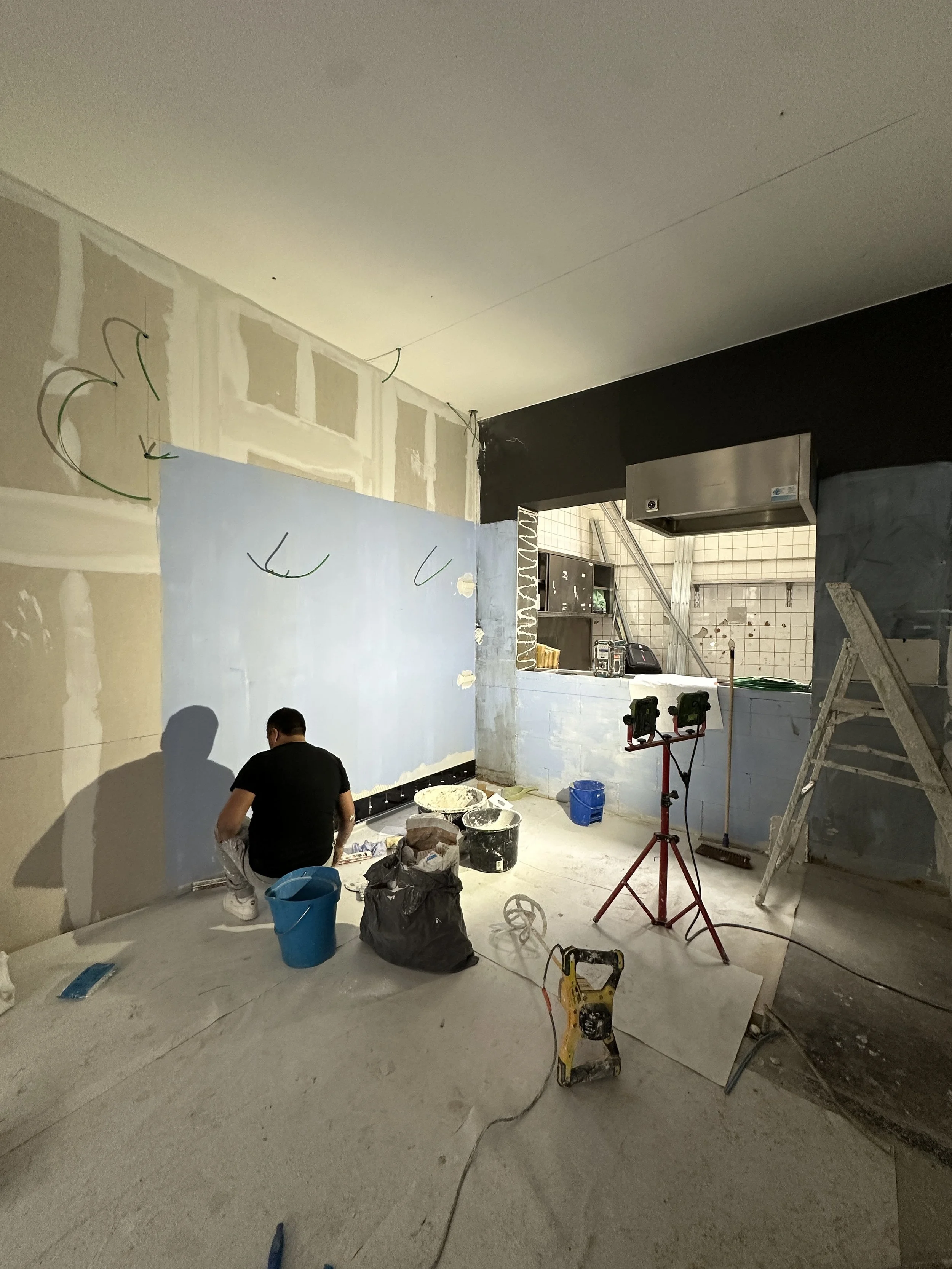 MANOS Build & Maintenance PANAM Kebab working site
A person working on a construction or renovation project indoors, with various tools and supplies scattered around, including buckets, a ladder, and lights, in a room with partially finished walls.