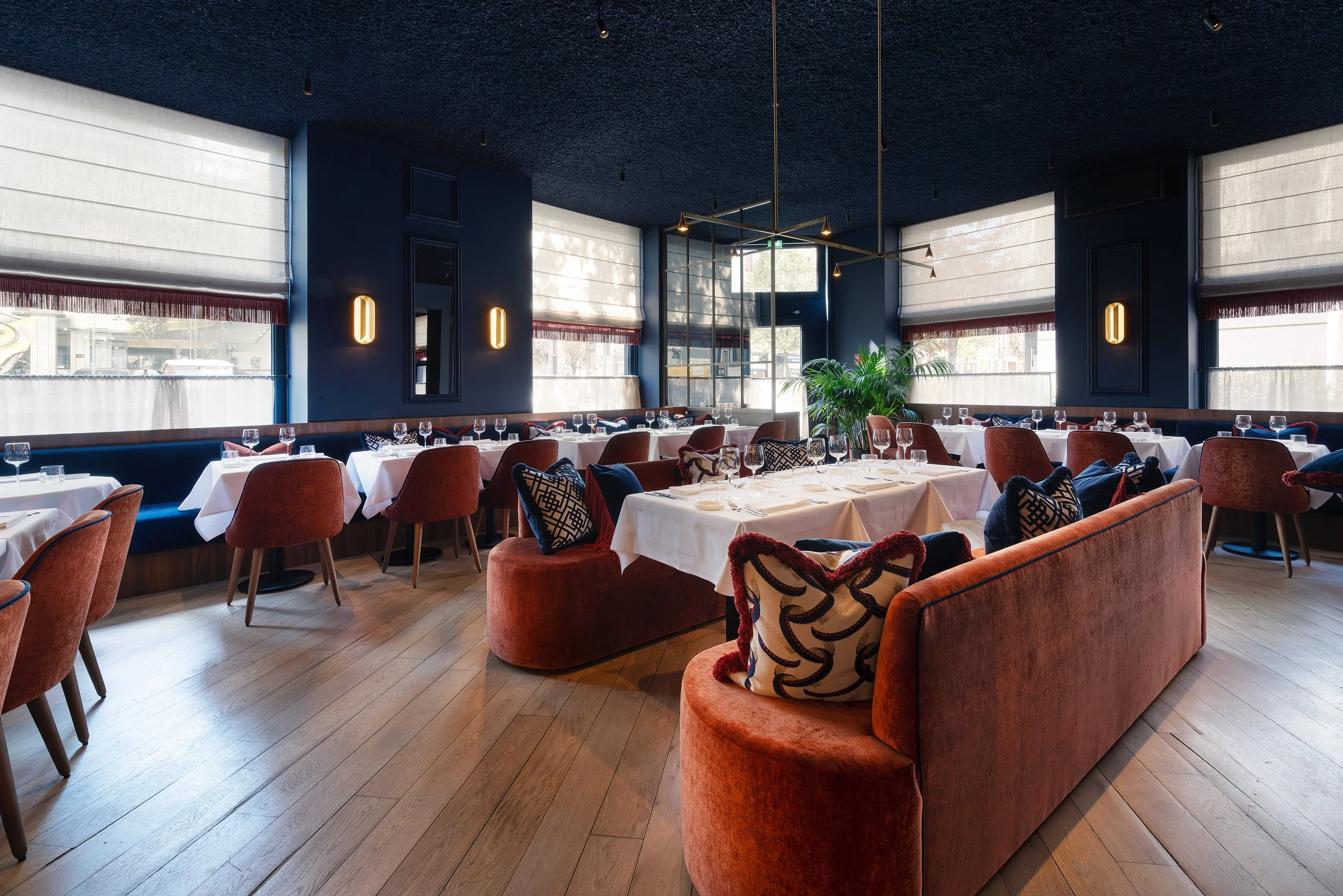 MANOS Build & Maintenance MUST 
Elegant restaurant interior with white-draped tables, plush reddish-brown chairs, decorative pillows, large windows with white blinds, blue walls with vertical lighting fixtures, wood flooring, and a large green plant.