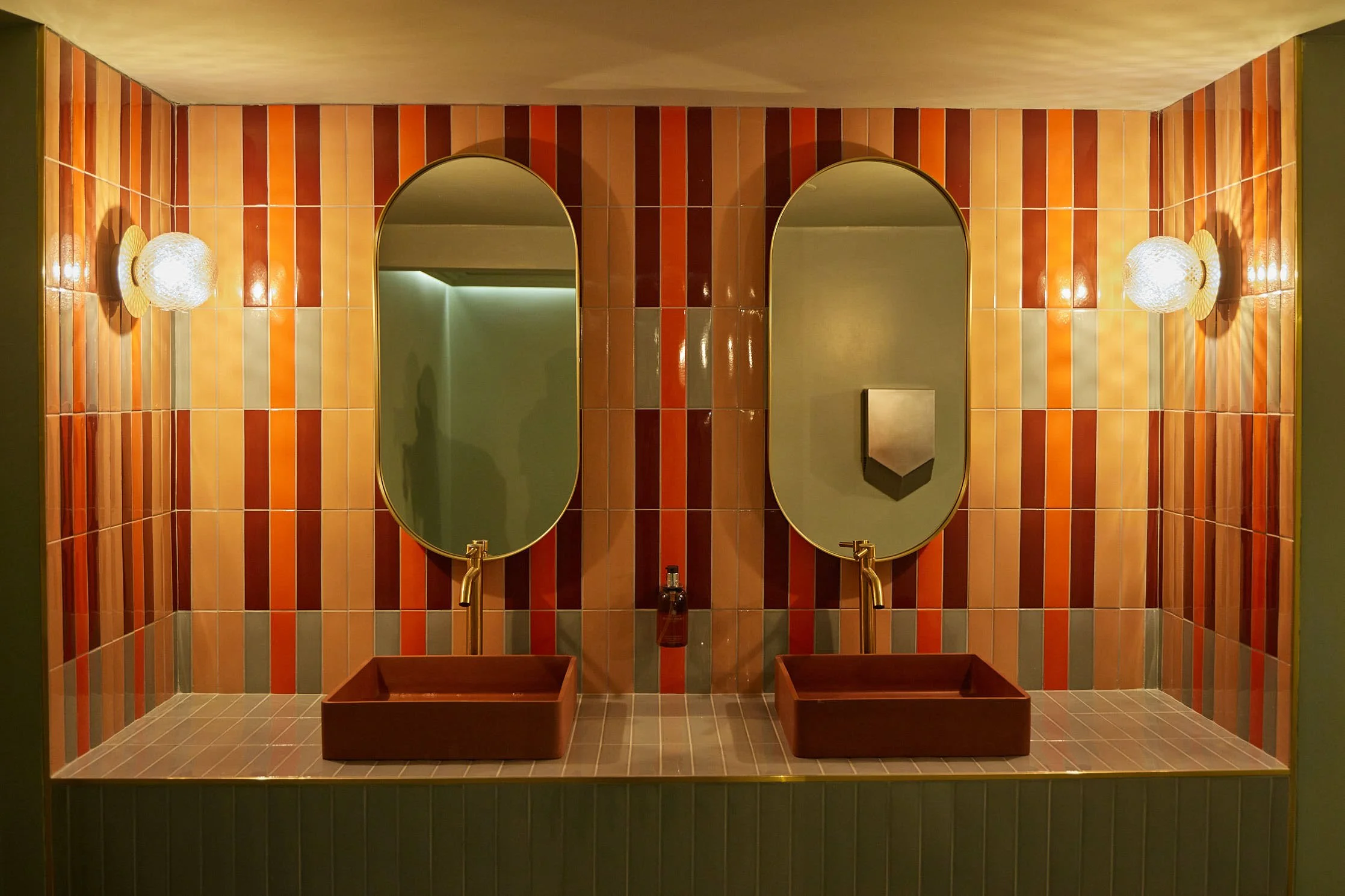 MANOS Build & Maintenance MUST restaurant working site
Double sink bathroom with two oval mirrors, colorful tile wall, two wall sconces, green walls on sides, and a countertop with two square basins.