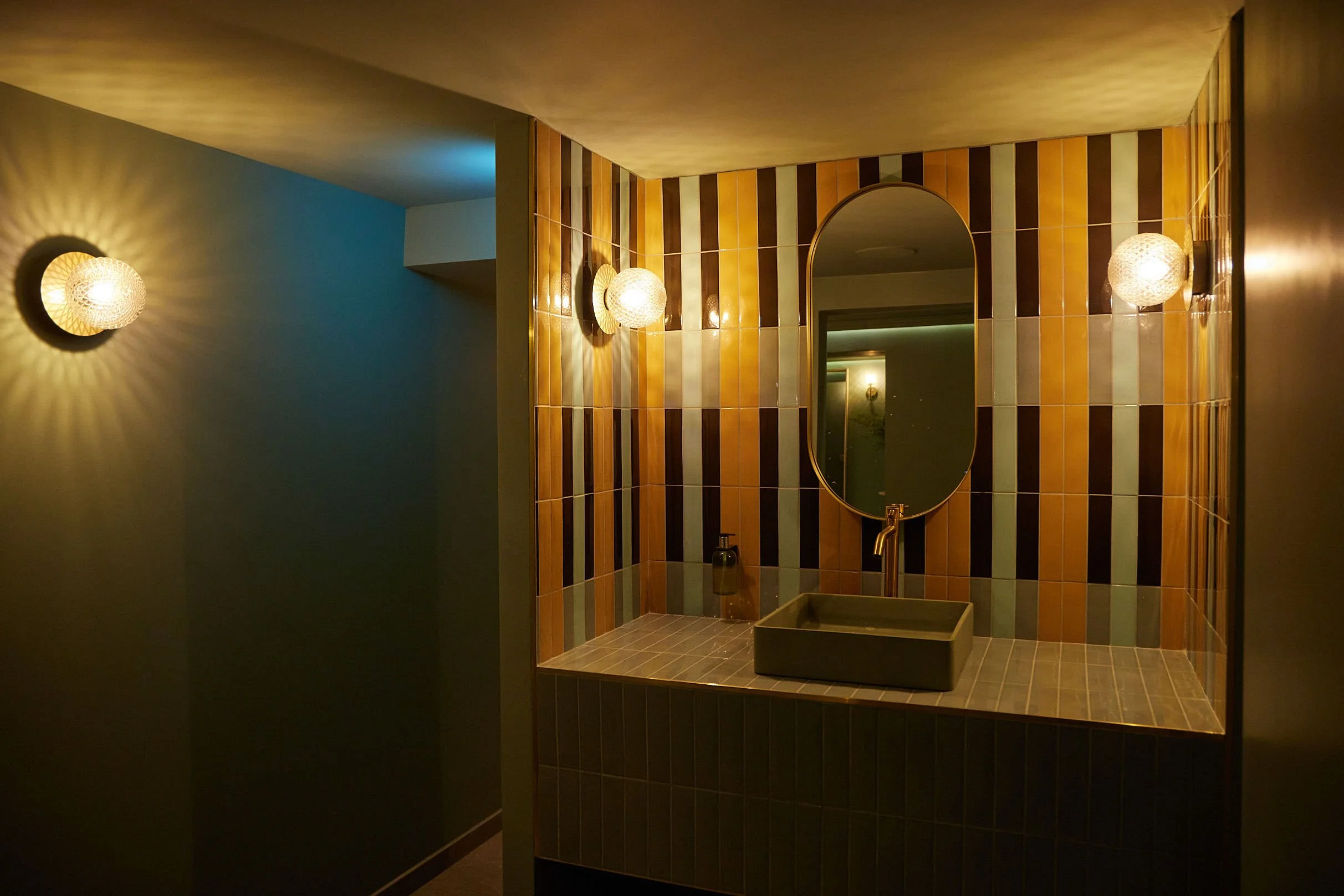 Bathroom vanity area with a rectangular sink, an oval mirror, and warm wall-mounted lights, decorated with vertically striped brown and beige tiles.