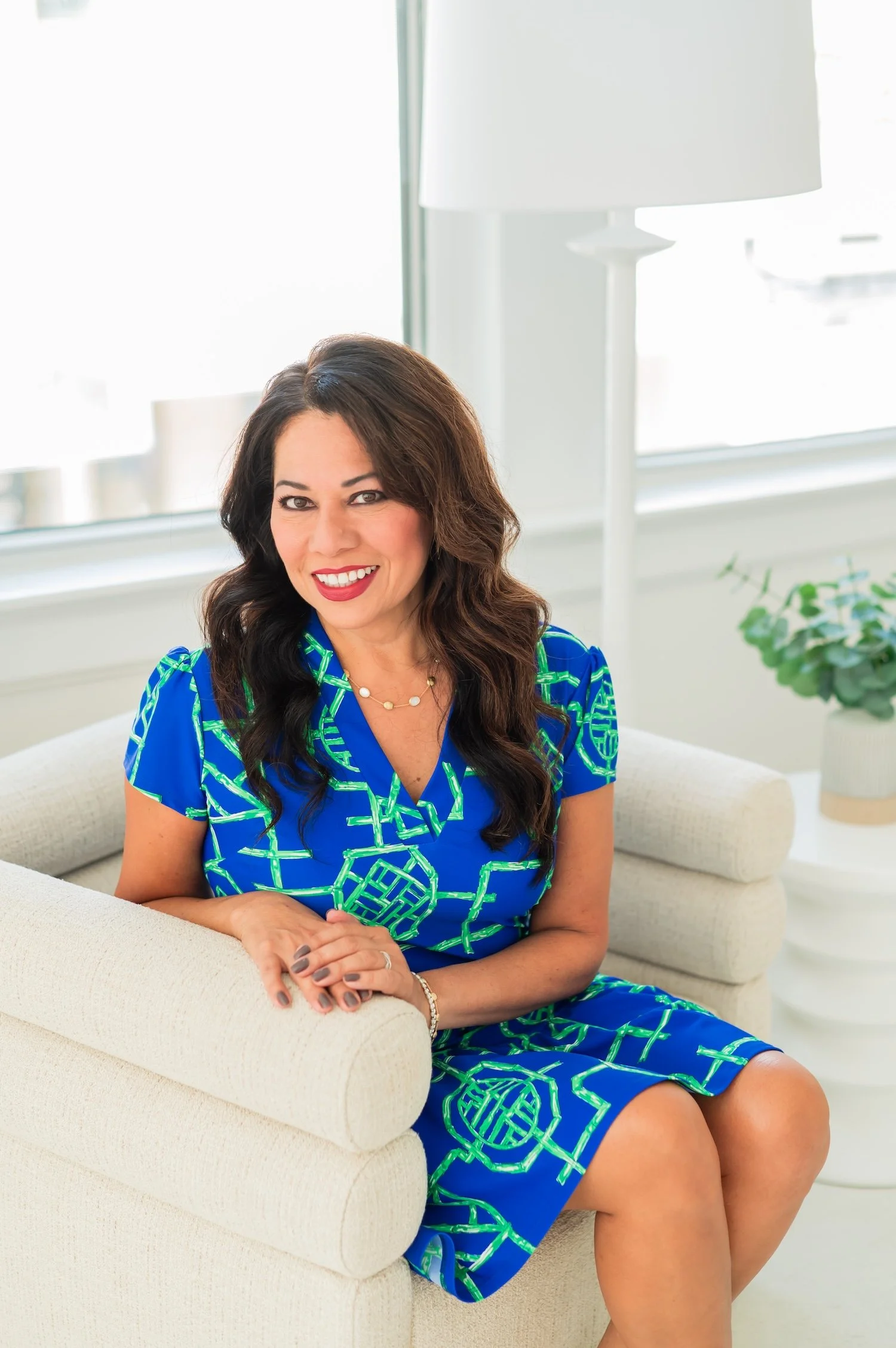 A woman with dark brown wavy hair, wearing a blue dress with green geometric patterns, sitting on a cream-colored couch in a bright room with a large window, a white lamp, and a potted plant in the background.