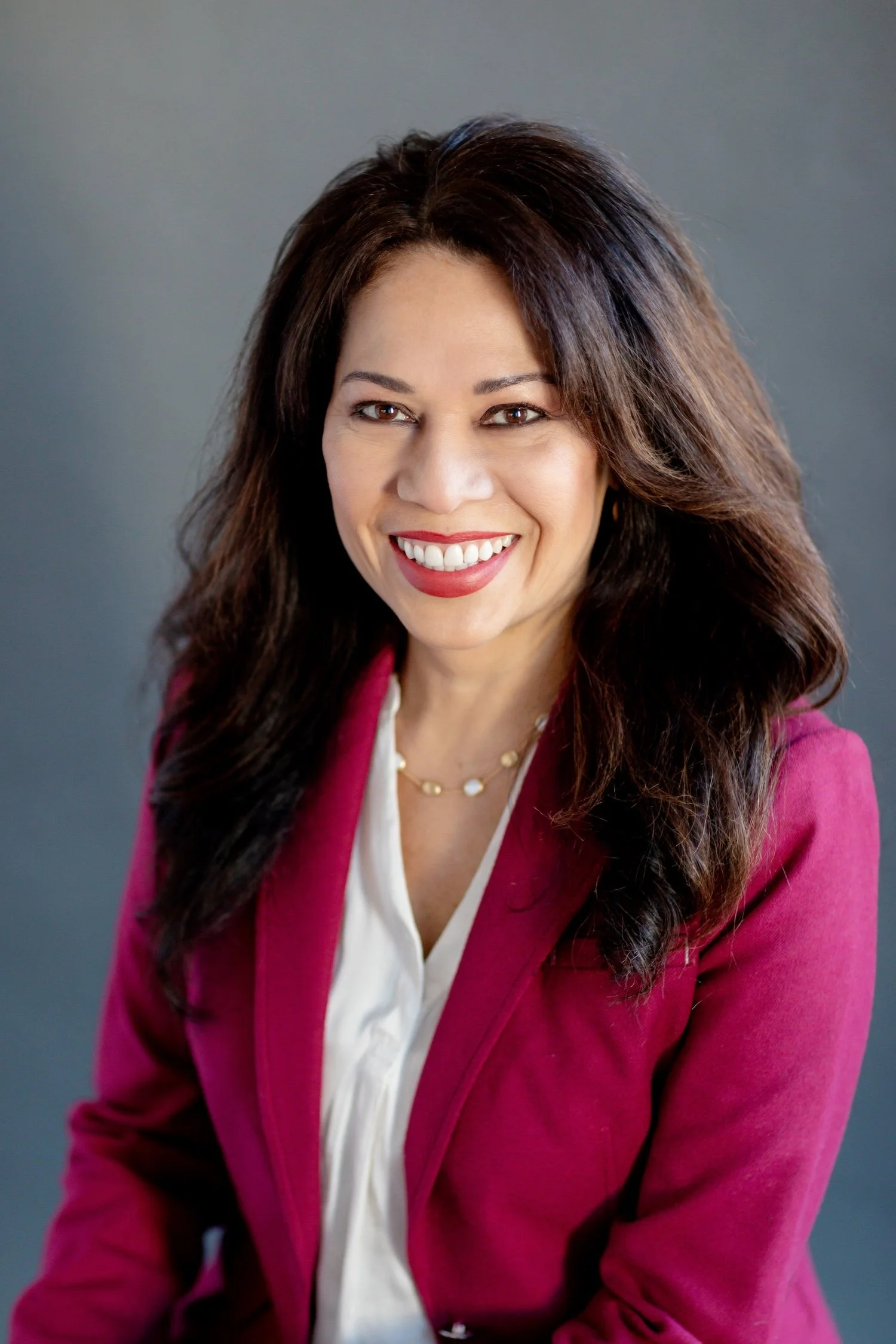 A smiling woman with long dark brown hair wearing a white blouse and pink blazer against a gray background.
