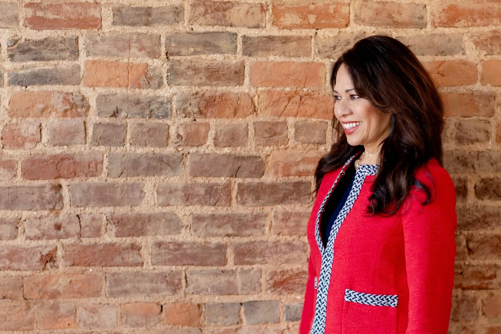 A woman with dark brown, wavy hair wearing a red blazer with navy and white patterned trim, standing against an exposed brick wall, smiling and looking to her left.