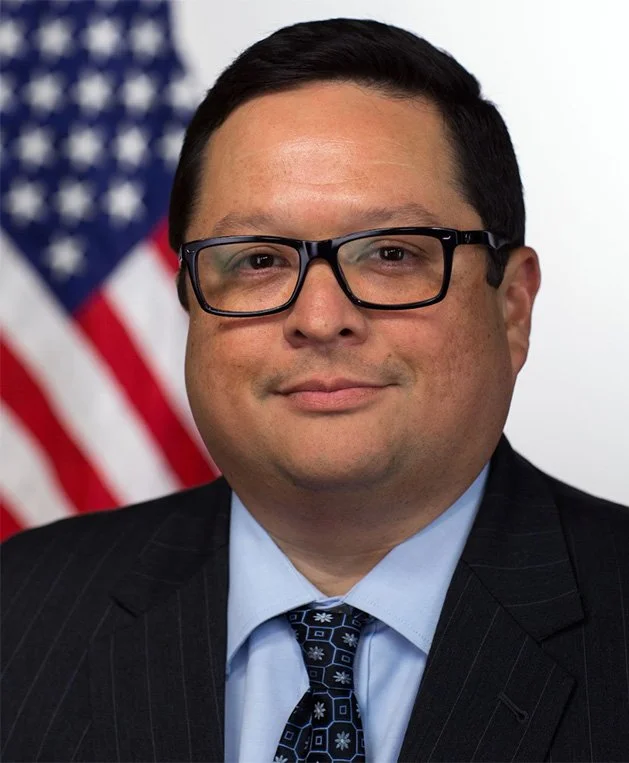 Portrait of a man with dark hair, glasses, wearing a suit and tie, standing in front of an American flag.