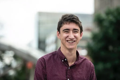 A young man smiling outdoors, wearing a maroon button-up shirt with small white polka dots, with city buildings and greenery in the background.