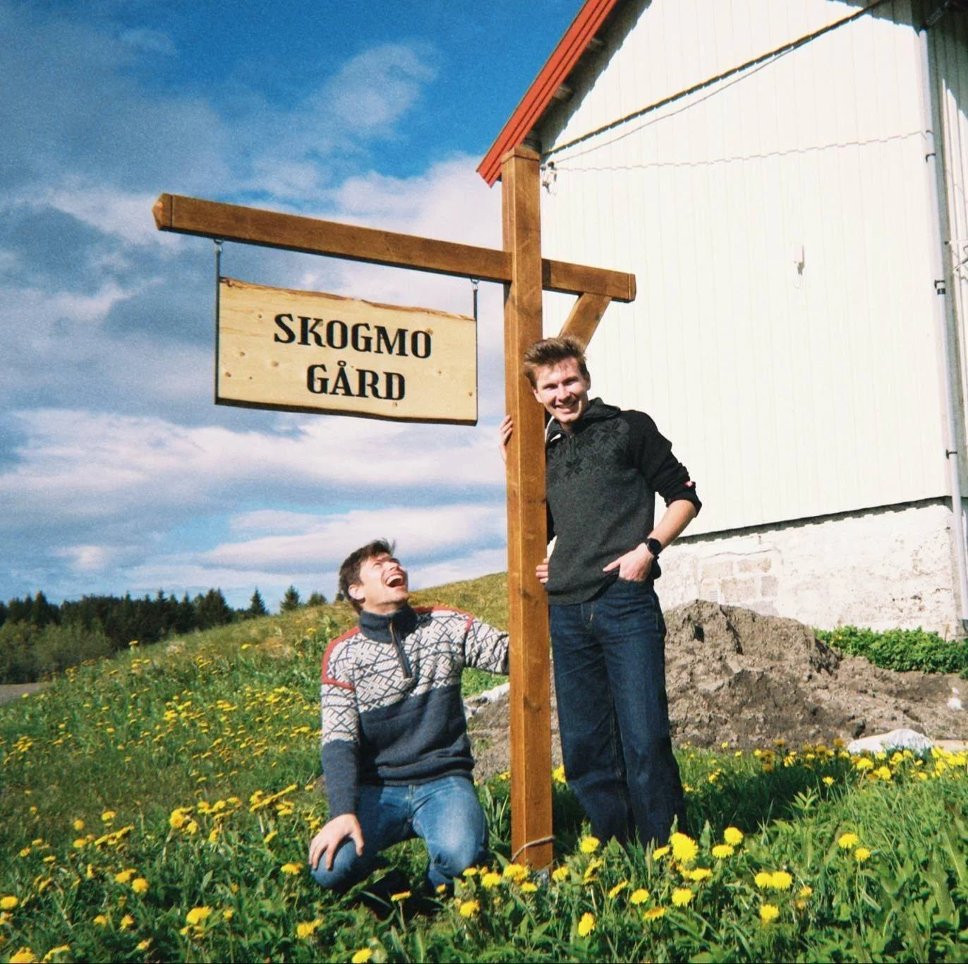 To unge menn ved en skilt som sier "Skogmo Gård" utenfor en bygning, omgitt av grønne marker med gule blomster, under en blå himmel med skyer.