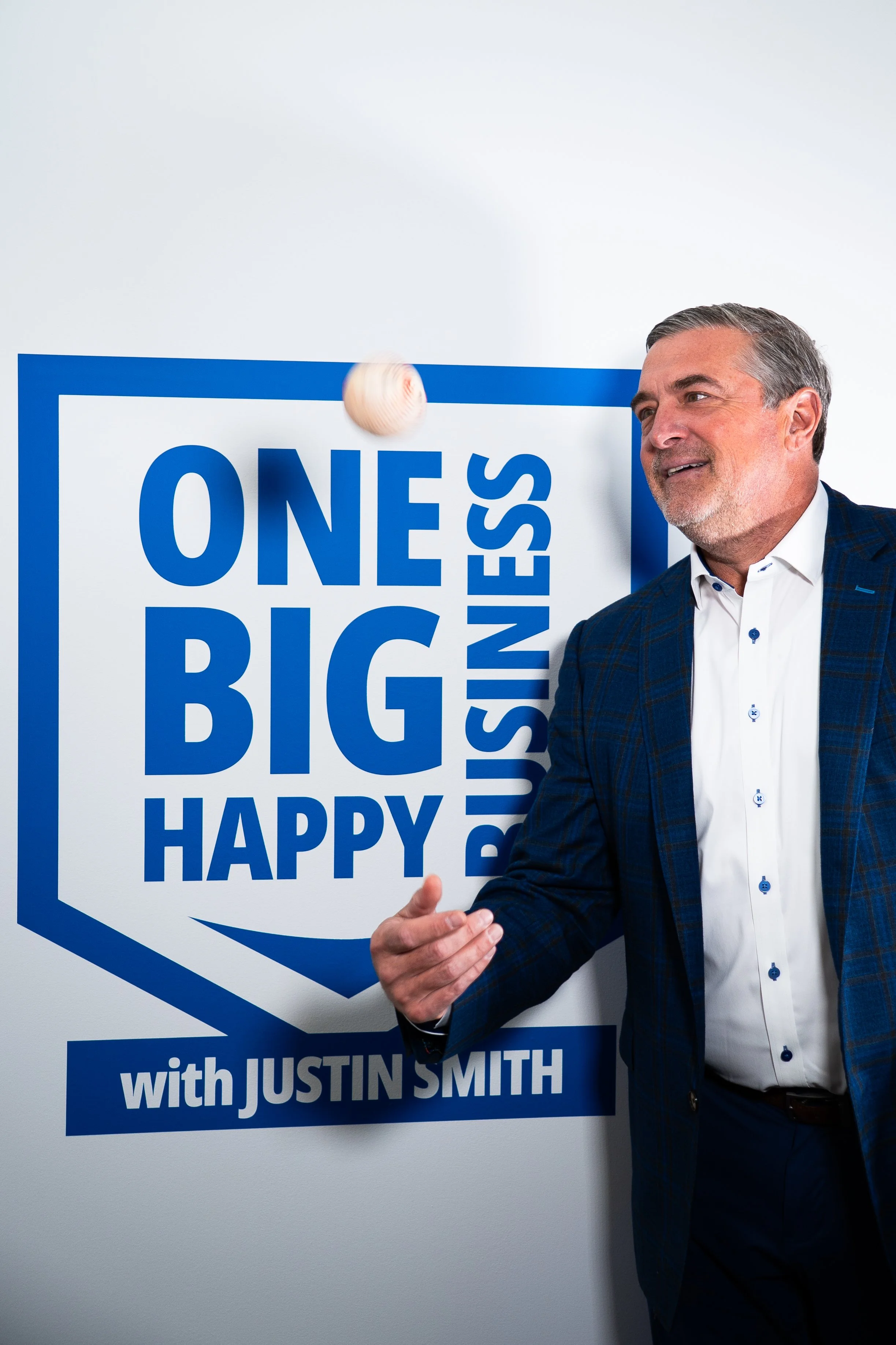 The host, in a suit, tossing a baseball in front of our studio sign that reads "One Big Happy Business with Justin Smith."
