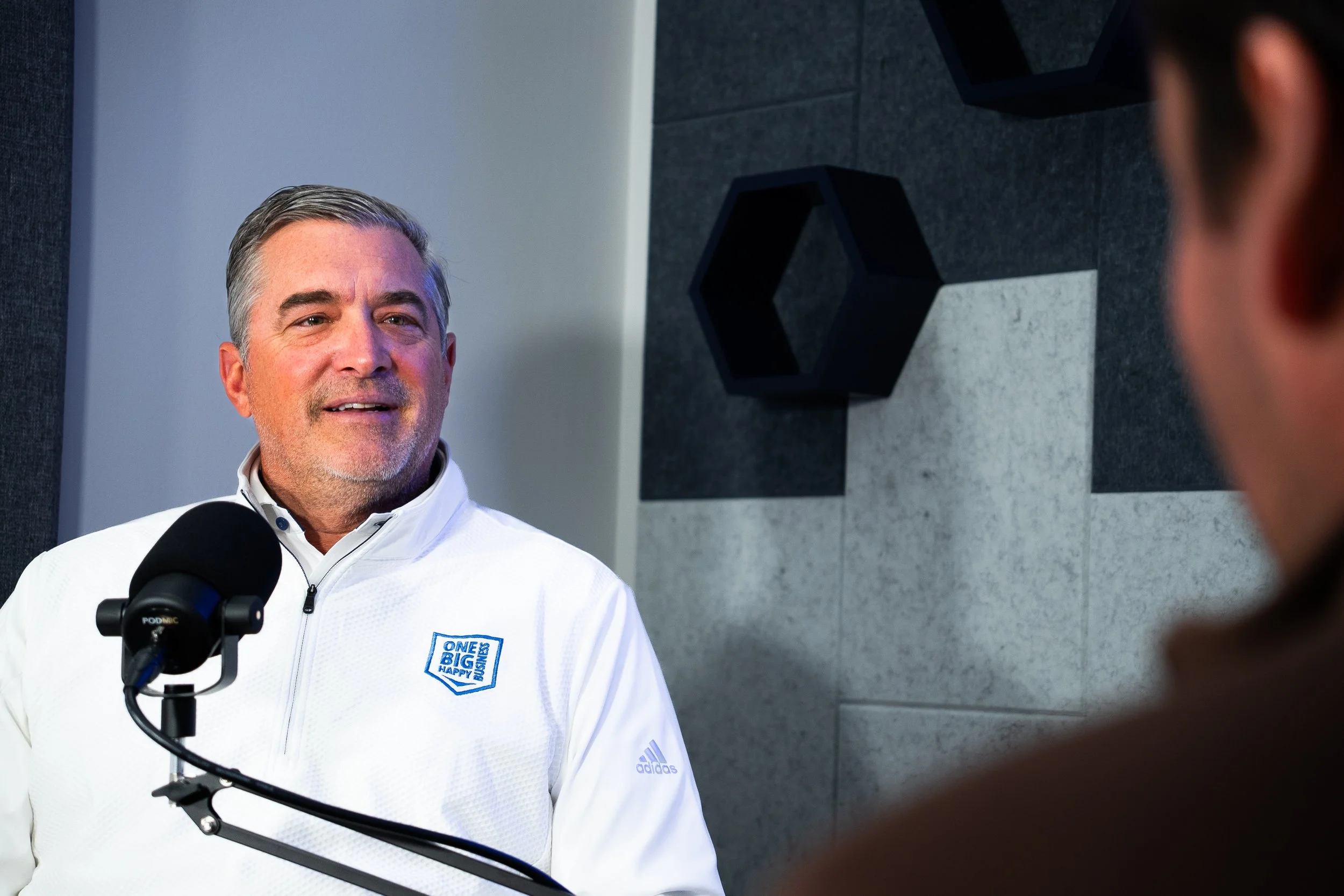 Justin Smith in a white One Big Happy Business jacket with a microphone in front of him, speaking during a podcast episode in our studio.