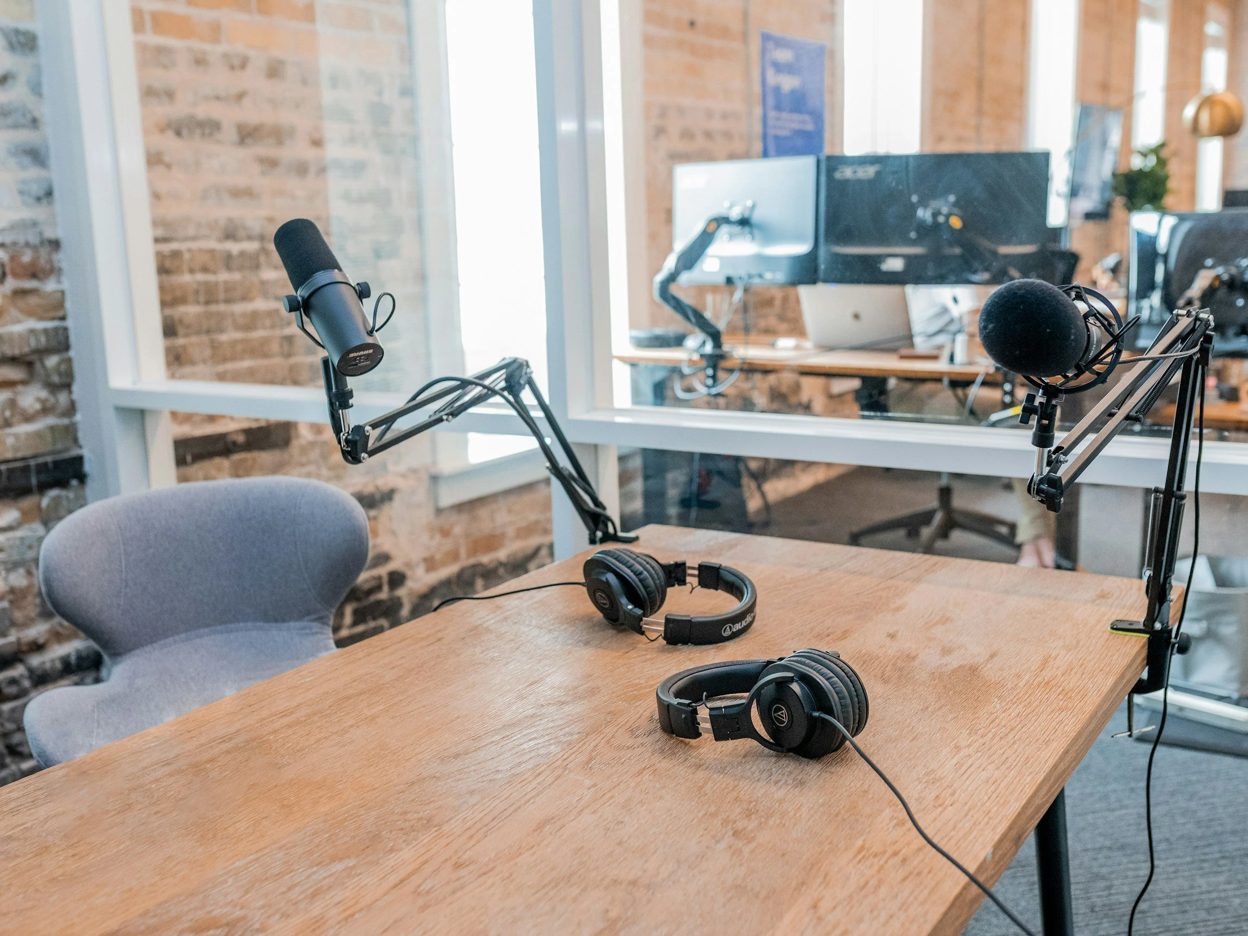 A podcast recording studio with two microphones on adjustable arms, headphones on a wooden table, and a computer monitor in the background. The room has brick walls and large windows.