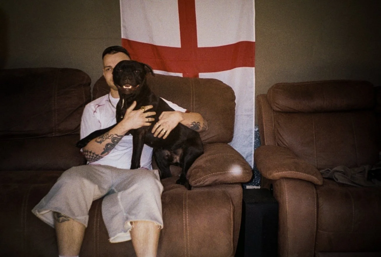 male fashion model wearing defender sportswear sat on a sofa with aa staffy dog and the England flag behind him