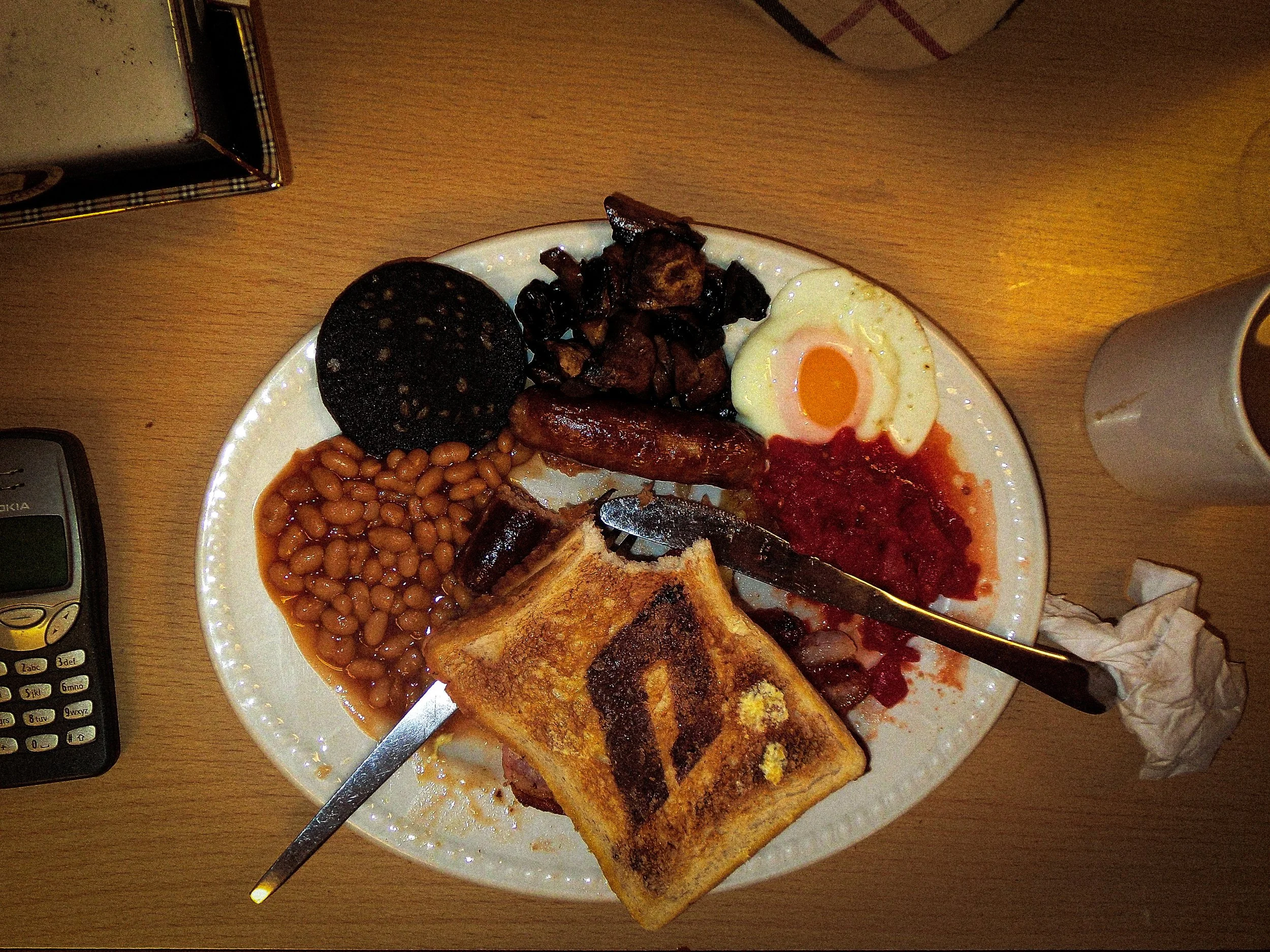 a  full English breakfast with 'D' Defenda menswear logo burnt into the toast, with a cup of tea next to the plate on one side and on the other a Nokia brick phone. Above the full English is a Burberry ashtray