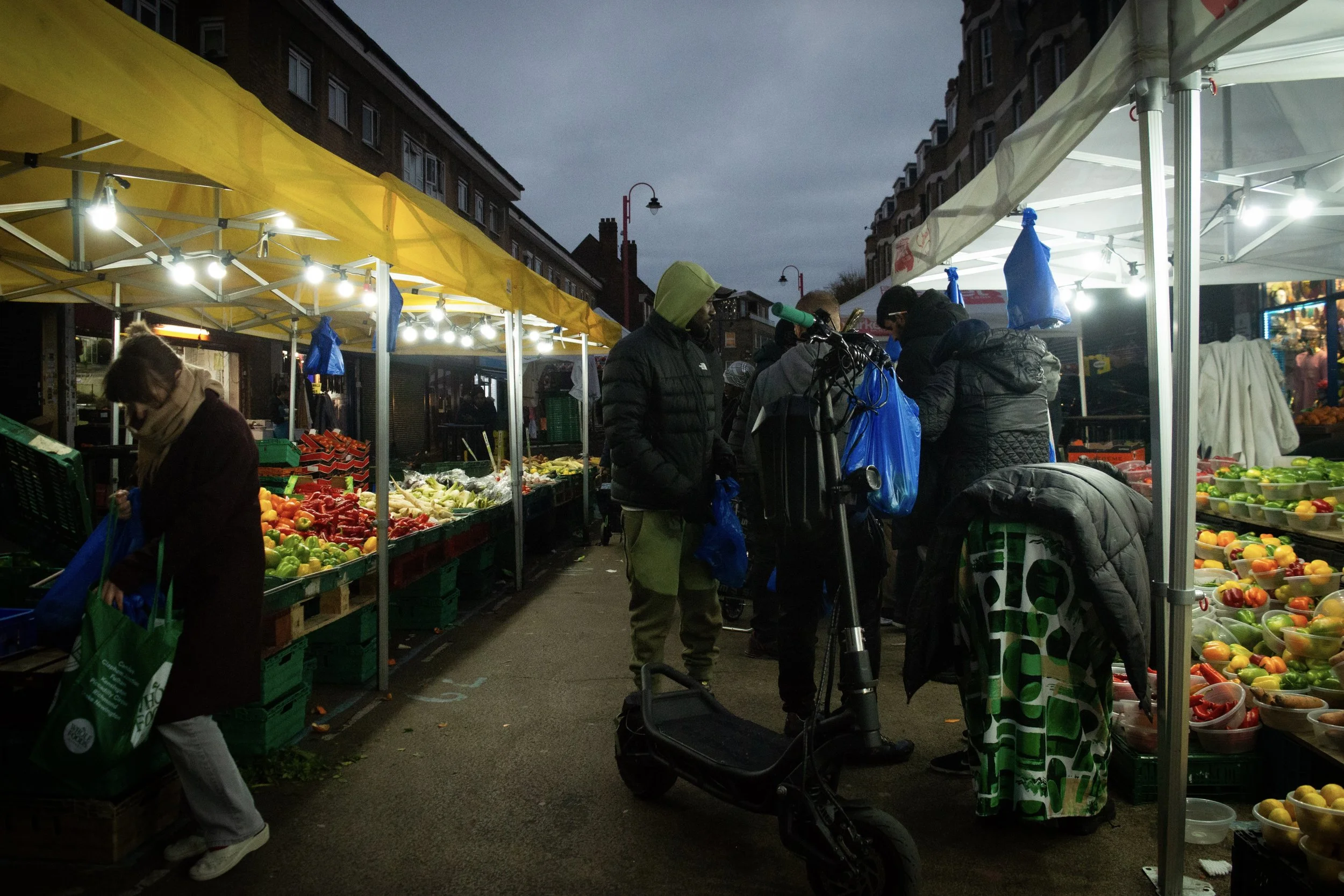 people shopping at a fruit and vegetable stall at East Street Market. In the middle of the isle is a young man wearing a tracksuit with an electric scooter