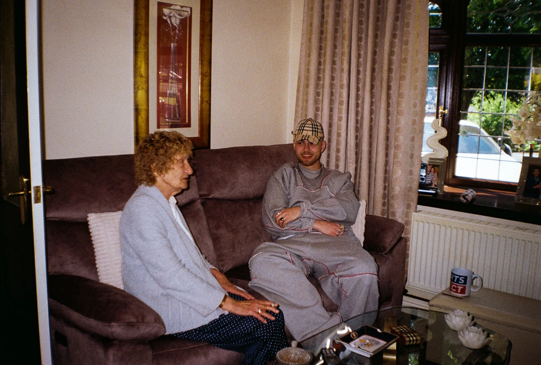 Model sitting with an elderly woman on a sofa on her house wearing defends ,ems wear tracksuit and Burberry cap for defenda mens wear fashion campaign 