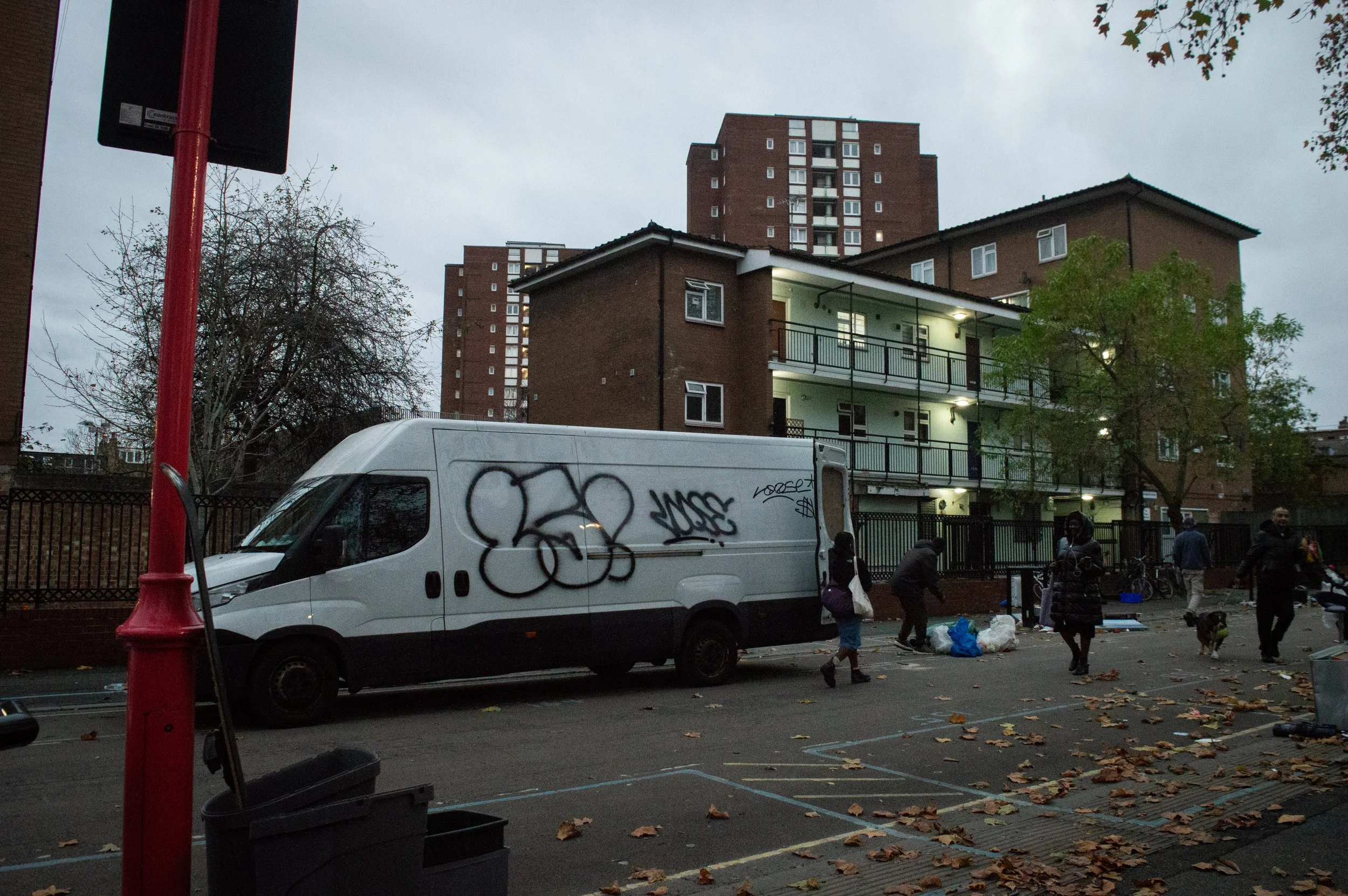 landscape of market workers packing up their stall at the end of the day into a white van with black graffiti on in front of a housing estate