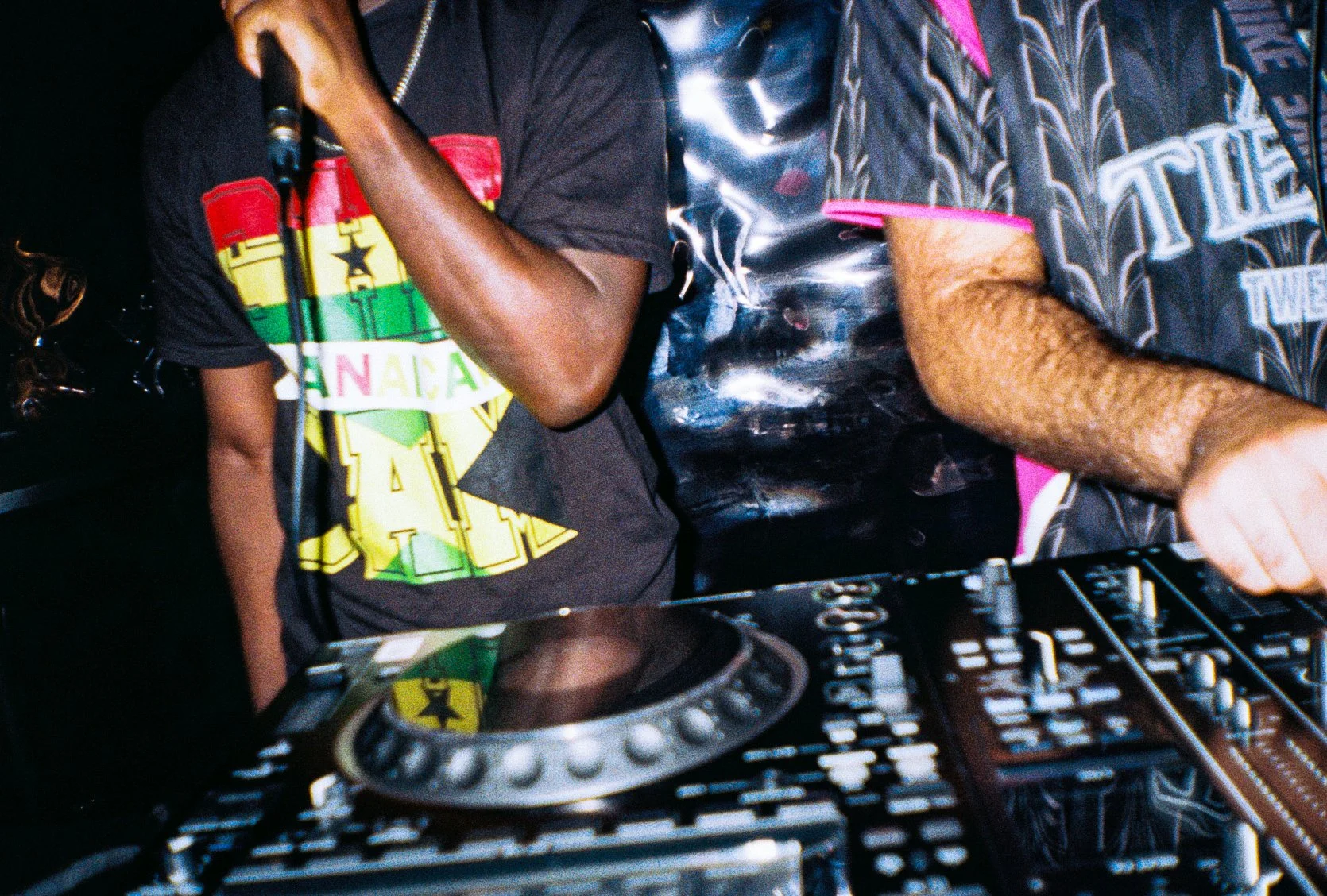 DJs mixing at a recorded grime set, with one person wearing a black T-shirt with a colorful Rasta-inspired design and another in a patterned black shirt with pink accents, with DJ equipment in front.