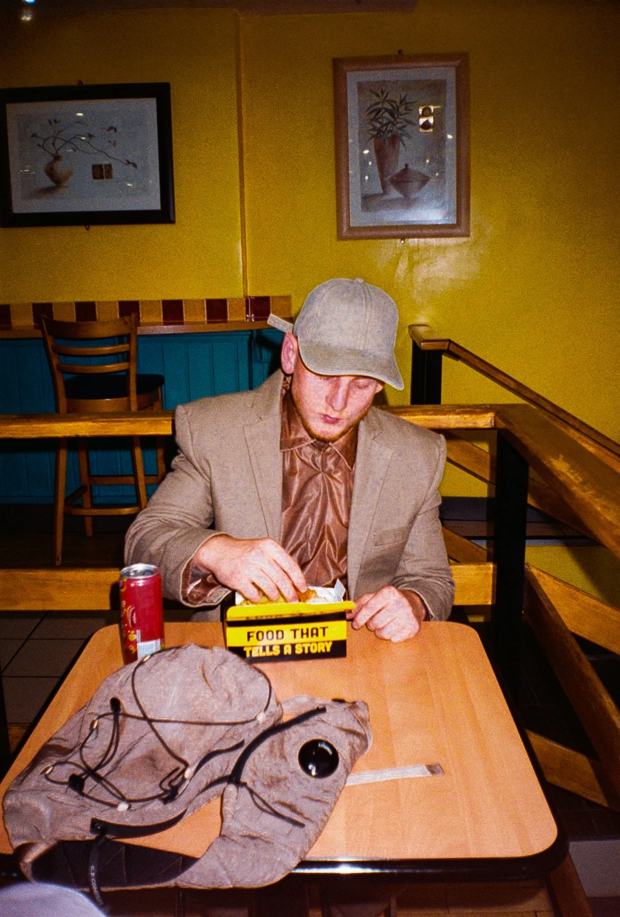 young man wearing beige/brown toned suit and a baseball style cap sat down eating chicken and chips in a chicken shop