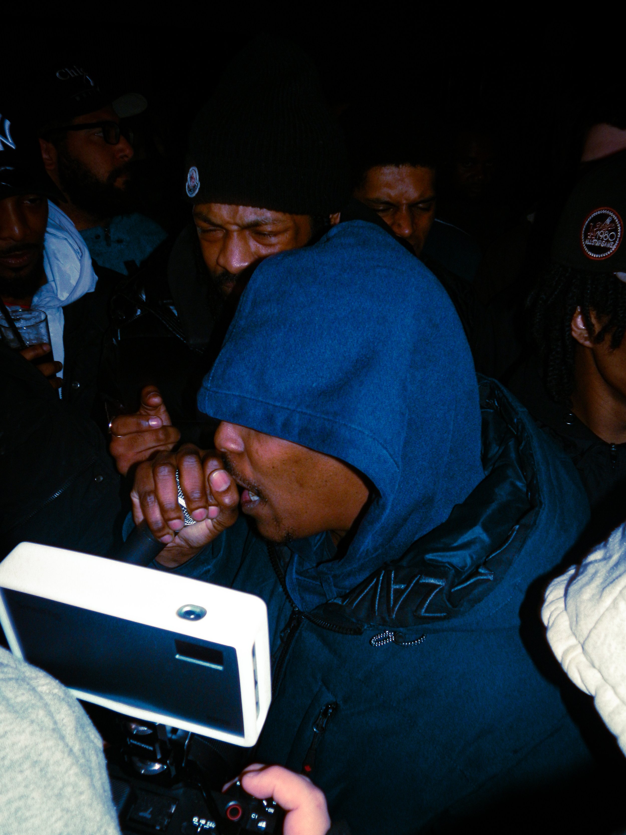 hooded mc performing at travspresents grime rave in the middle of a crowd with camcorders in his face