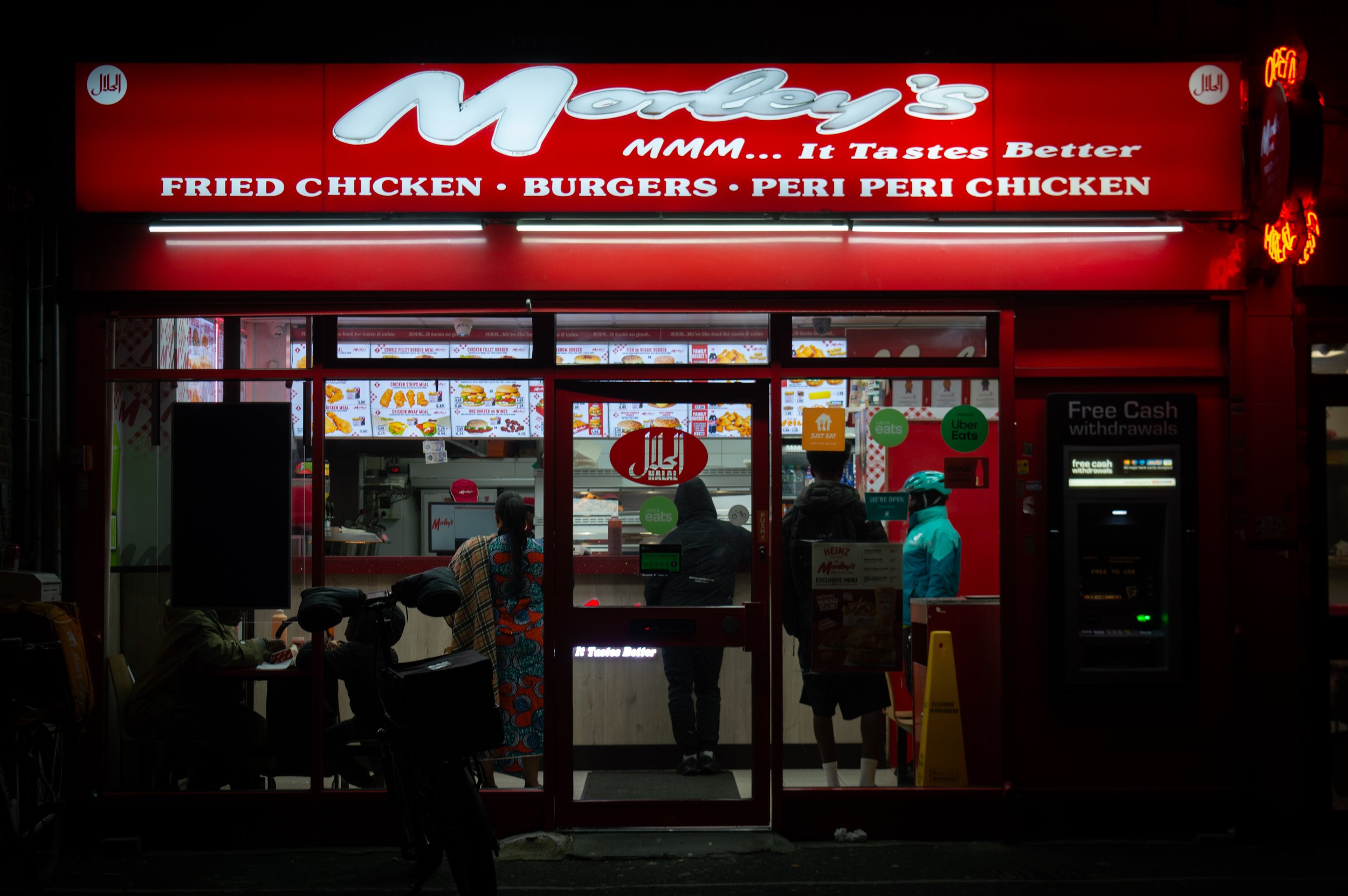 Front view of a fast-food restaurant named Morleys's with red signage, illuminated menu boards inside showing fried chicken, burgers, and peri peri chicken, signs for Uber Eats and Eat, and a cash withdrawal kiosk outside at night time