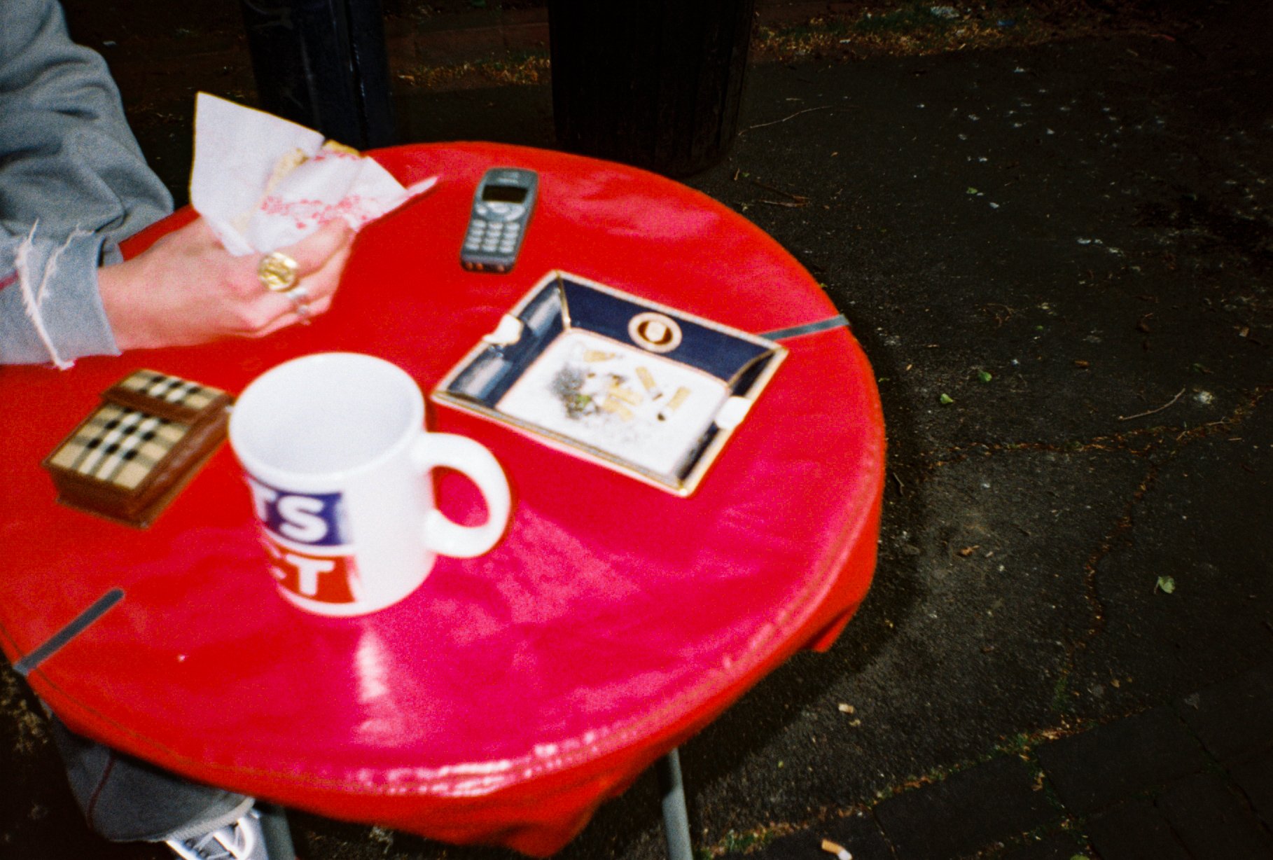 landscape detail photo of a burger van table, on the table is a sports direct mug, a Burberry ashtray, Burberry cigarette case, Nokia phone, and a mans hand holding a burger