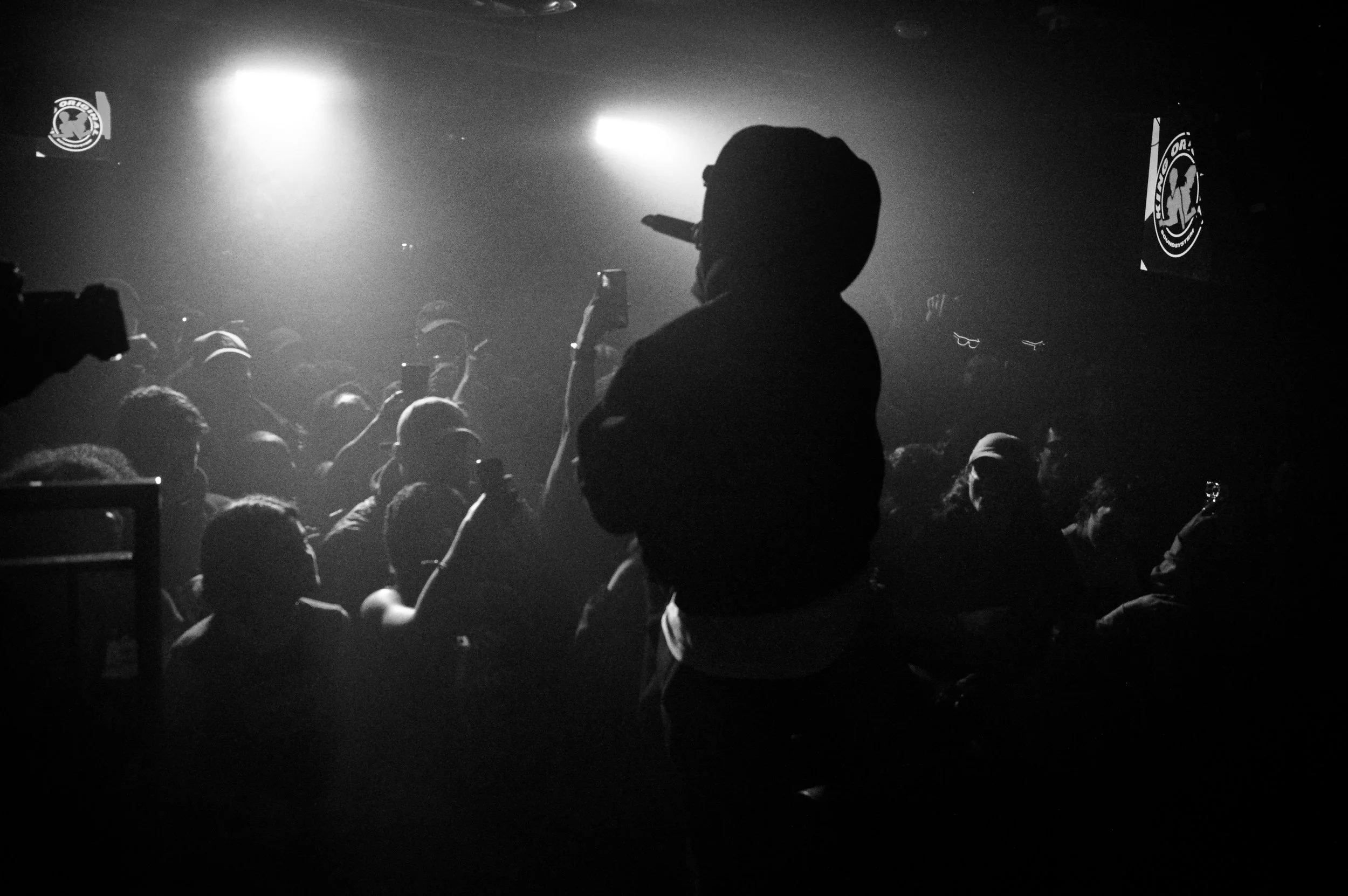 A black-and-white photo of a crowd at a rave, with a person in the foreground taking a photo or video using a smartphone.