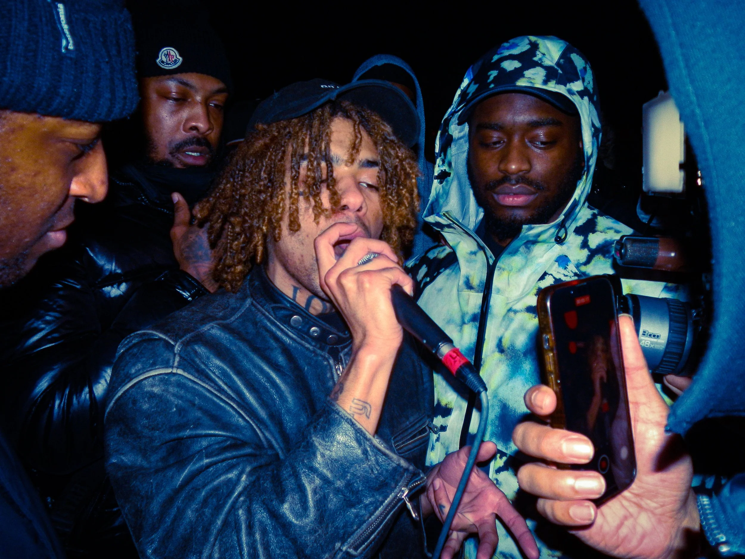 grime musician SBK performing into a mic and a camera with mcs faultzy, elf, and renz standing around him at a travspresents rave