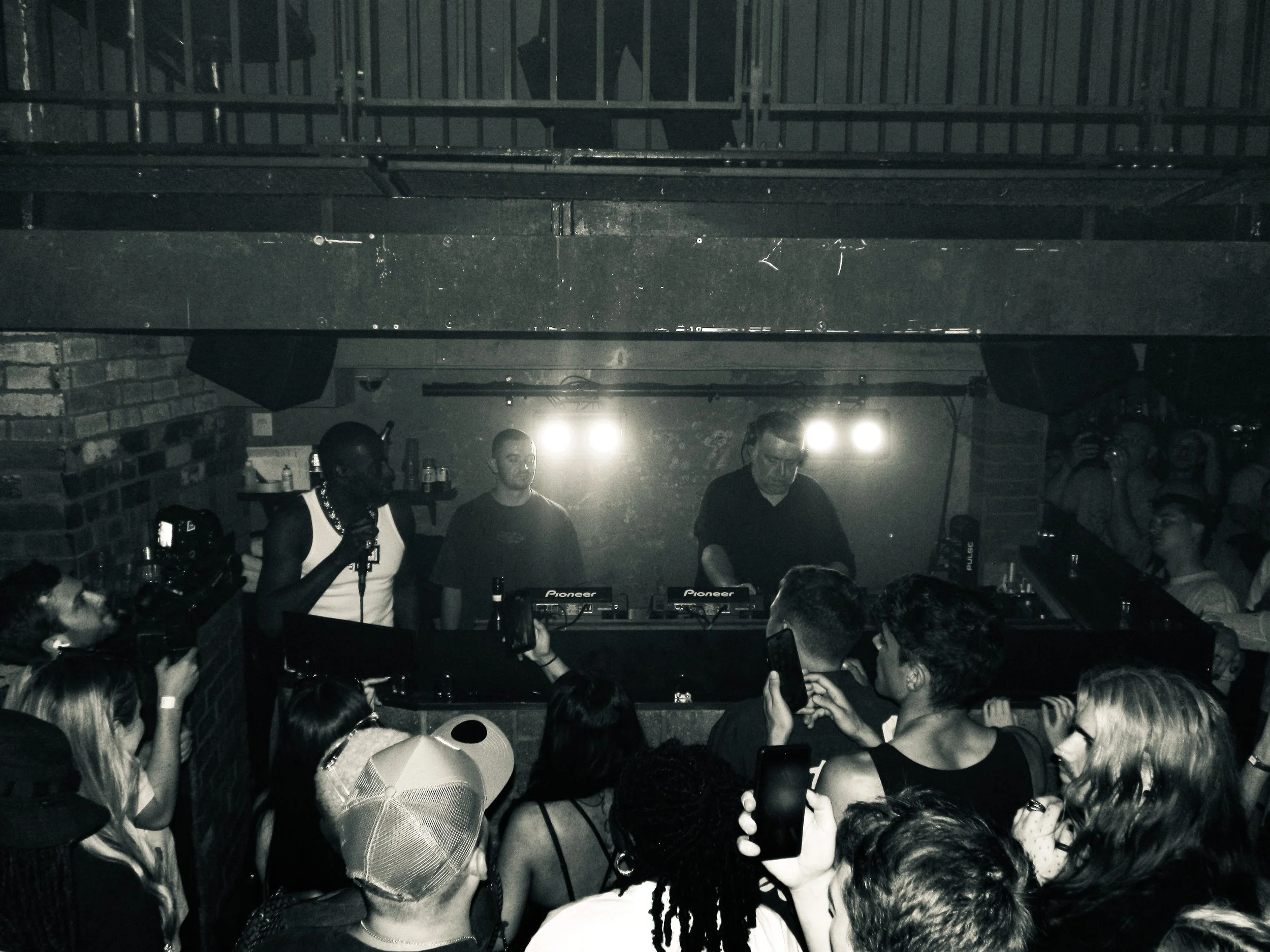 DJ slimzee performance at a nightclub with a crowd dancing and taking photos, black-and-white photo showing a DJ behind a mixer, and people enjoying the music at a grime rave 