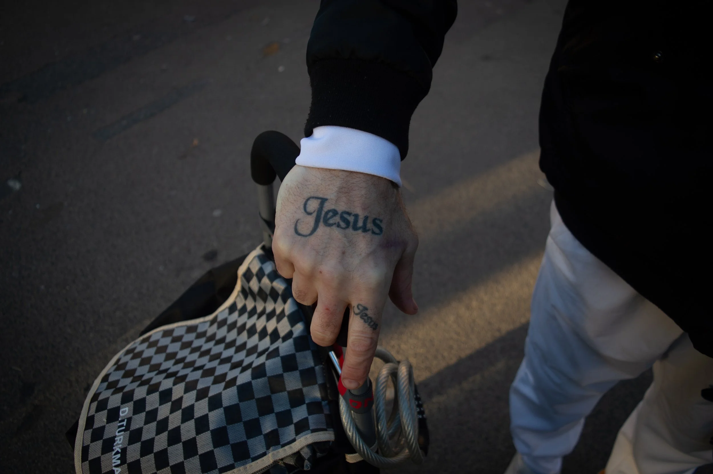 an elderly mans hand resting on the handle of his checkered shopping trolley with 'Jesus' tattooed across it.