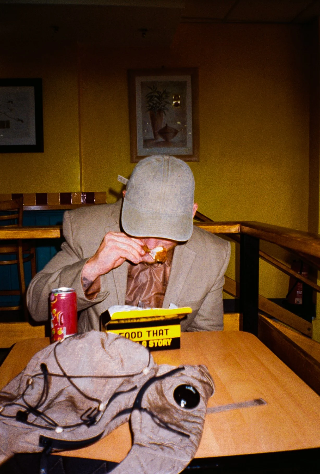 young man wearing beige/brown toned suit and a baseball style cap sat down eating chicken and chips in a chicken shop