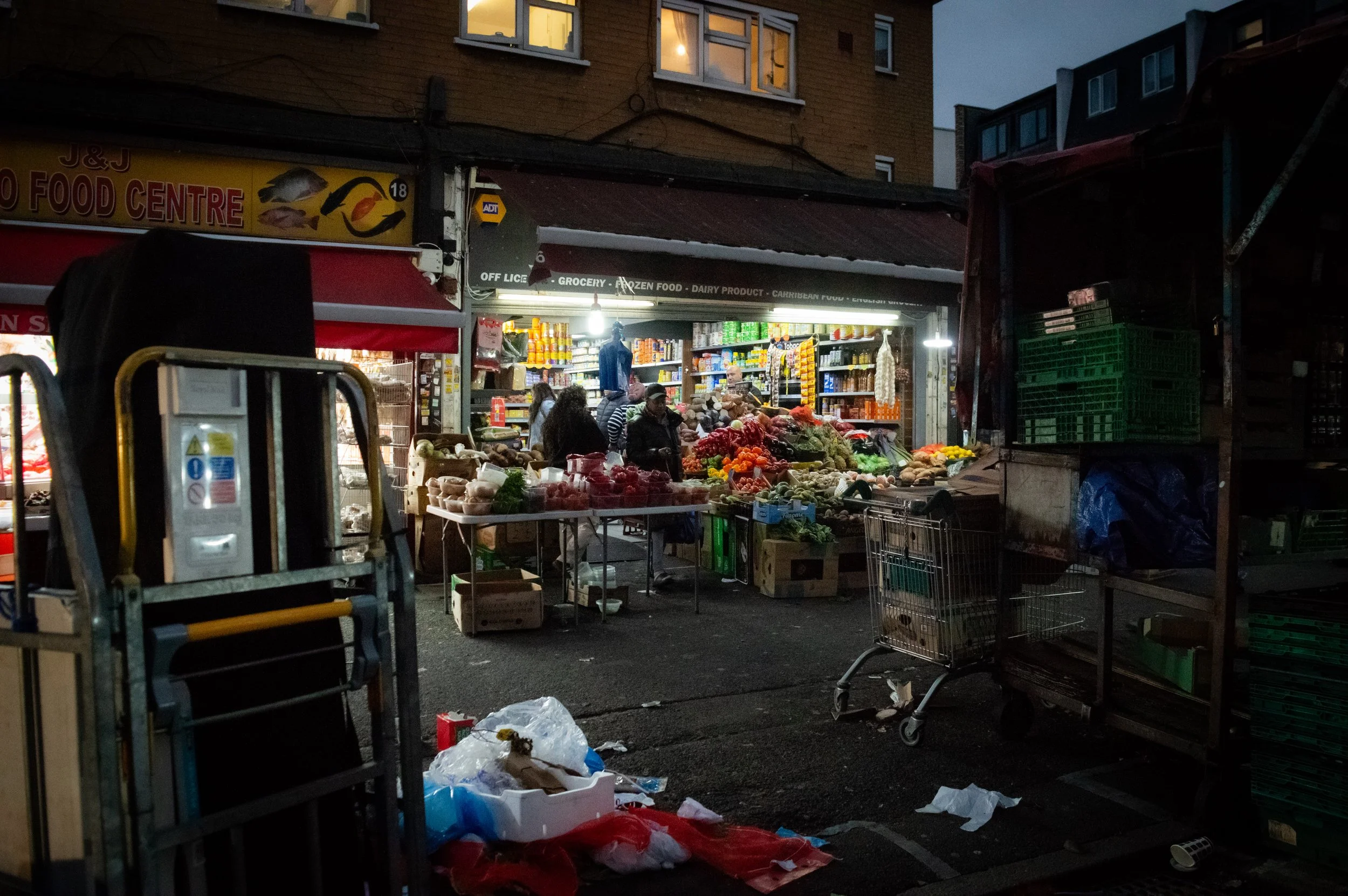 evening landscape of the outside of an indoor market style ethnic food shop with customers inside and walking past and some rubbish on the streets