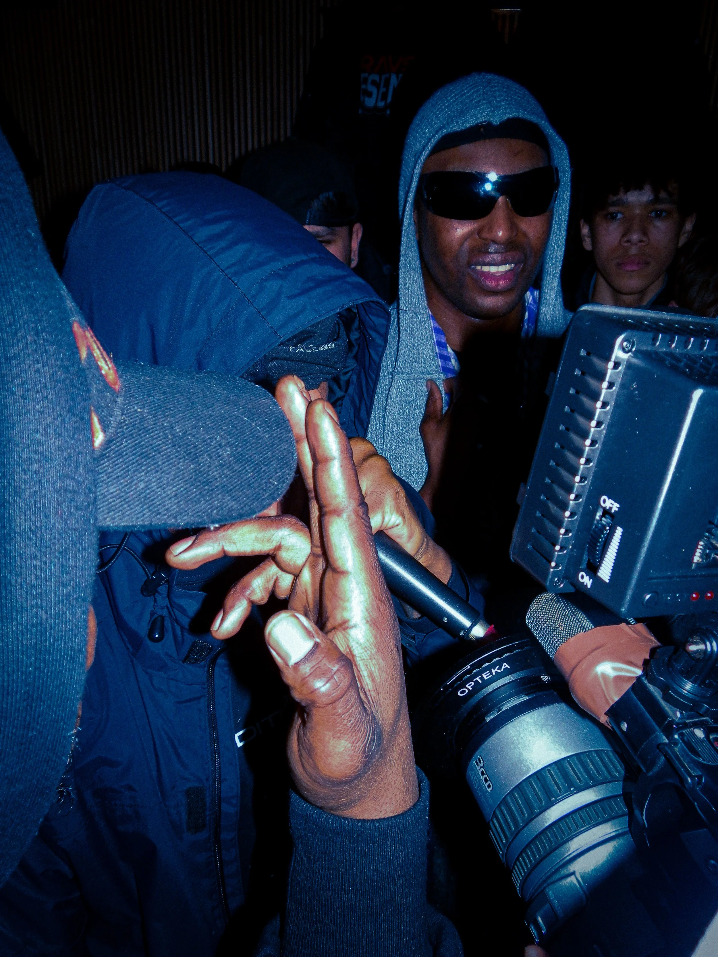 Grime mc merry ace performing into mic and cam corders with someone holding up gun fingers covering his face 