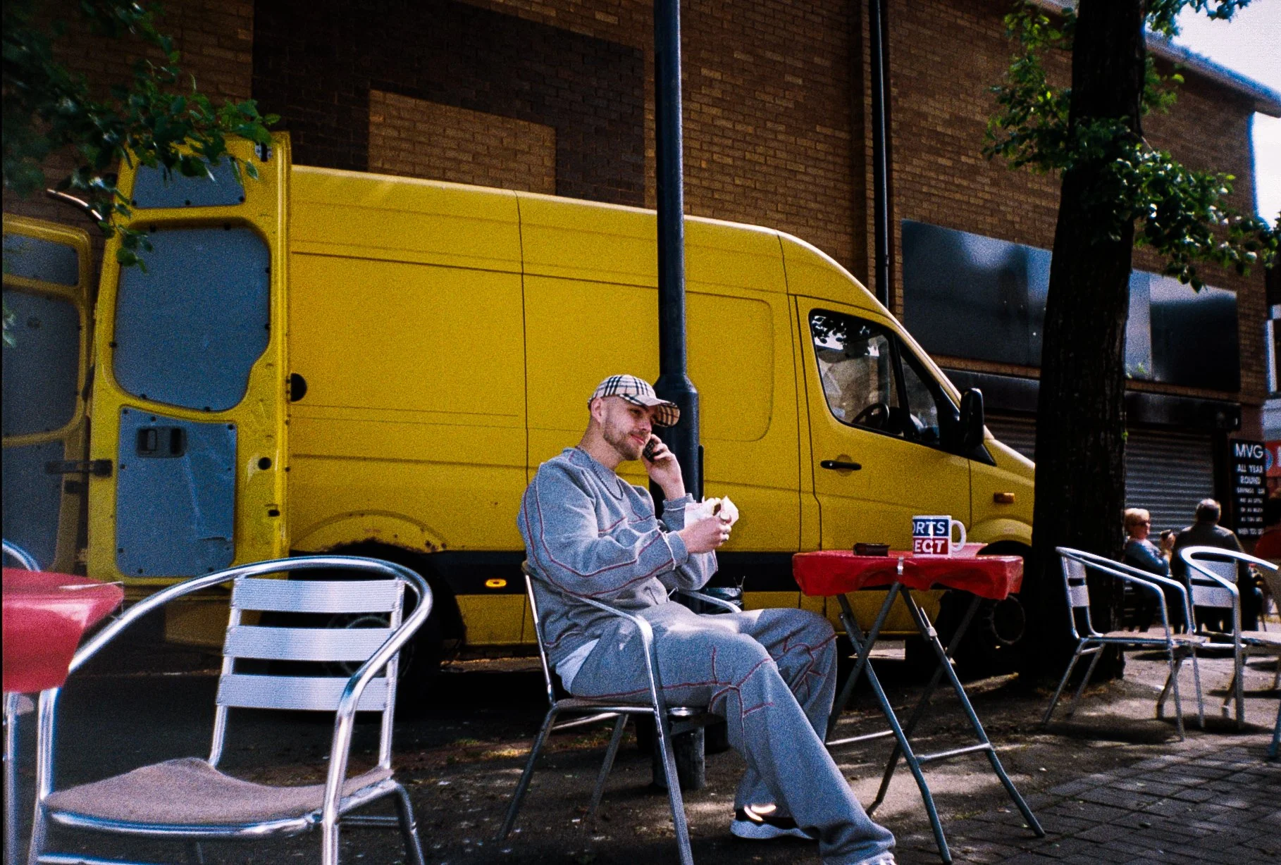 Male fashion model on the phone sat at a burger van table eating a burger, yellow van in the background