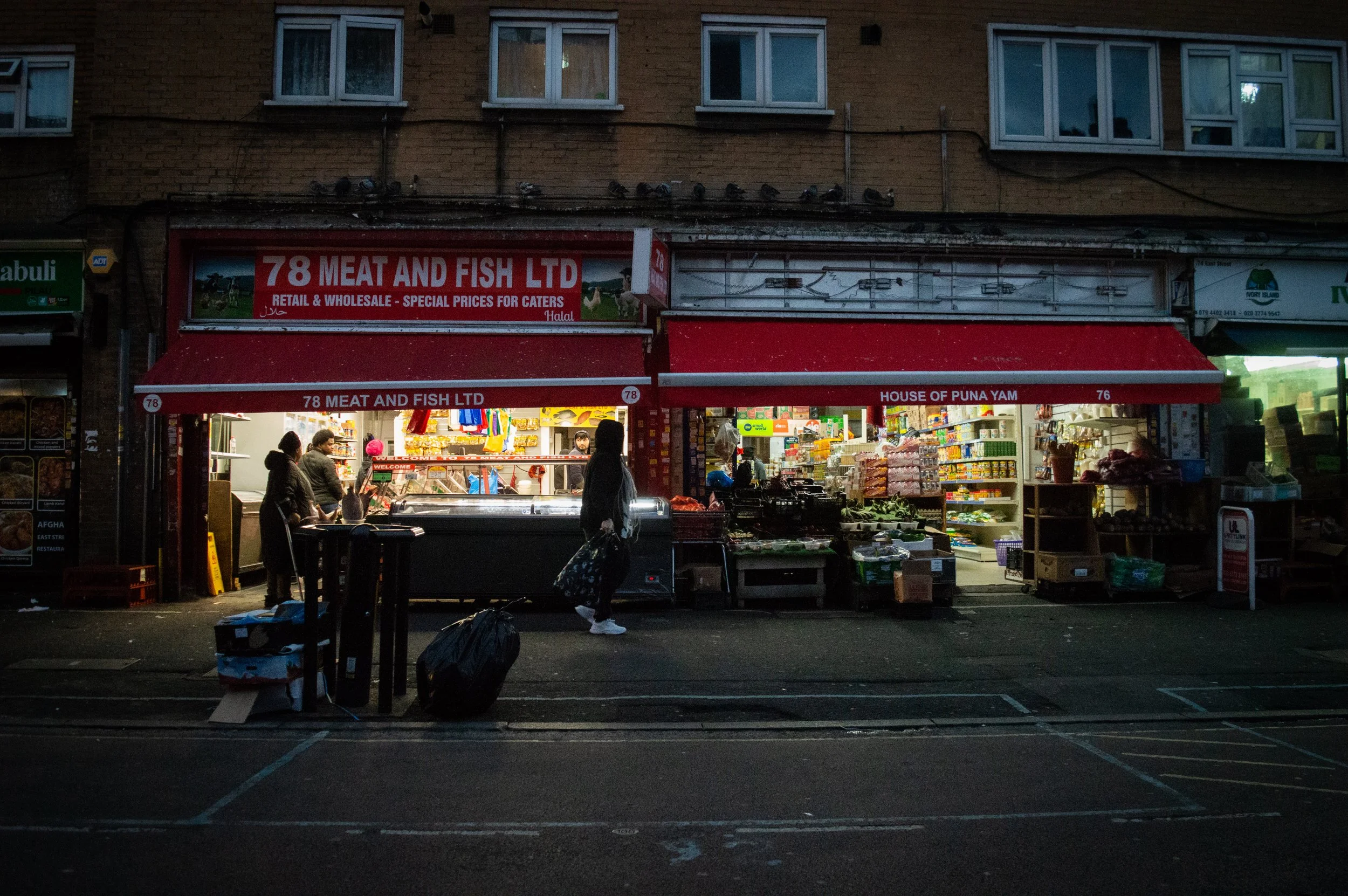 evening landscape of the outside of an indoor market style ethnic food shop with customers inside and walking past and some rubbish on the streets