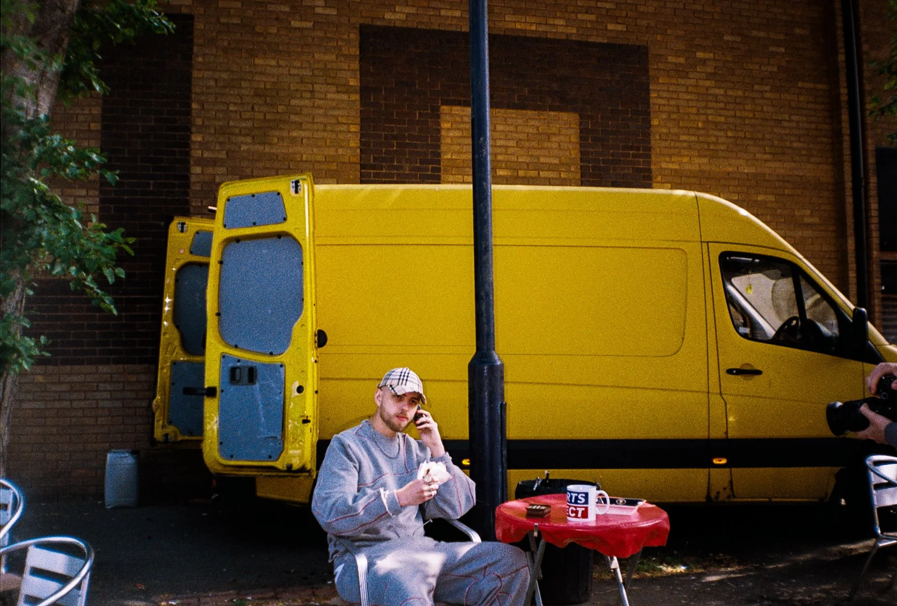Male fashion model on the phone sat at a burger van table eating a burger, yellow van in the background