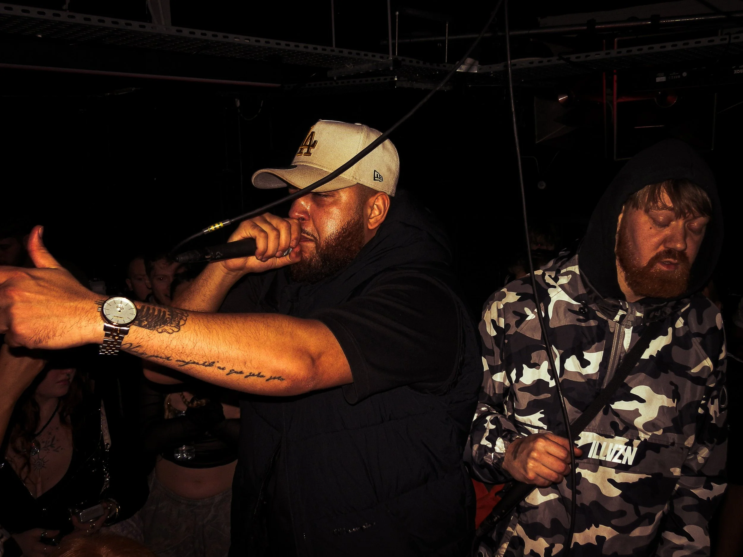 Two men performing on stage at a rave, one mcing into a microphone wearing a baseball cap and the other in a camouflage hoodie holding a microphone.